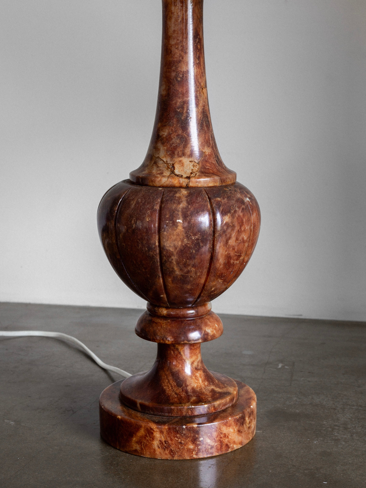 A close-up view of the base of the Large Italian Alabaster Stone Lamp by Spigel, showcasing its polished, brown marble finish with a rounded, ribbed design that exudes a neoclassical style. The lamp's electrical cord is partially visible on the left side, resting on a flat, concrete surface. The background is a plain, light-colored wall.