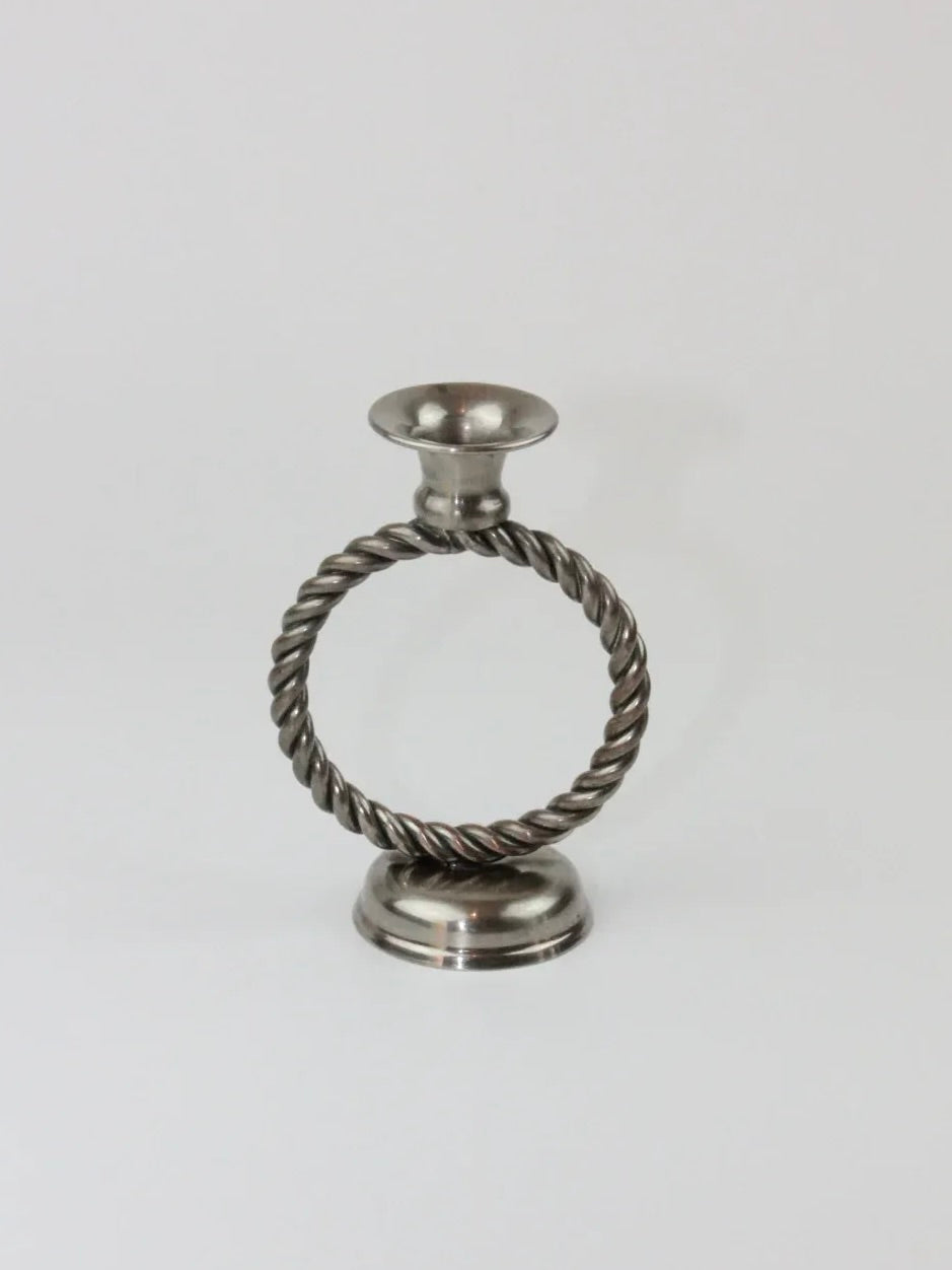 The Mental Candle Holders from Boga Avante Shop feature a circular, twisted rope-like design in the center of a metallic structure. The round and sturdy base supports a wide, shallow dish perfect for holding a candle. This piece exudes a classic and sophisticated look against the plain white background.