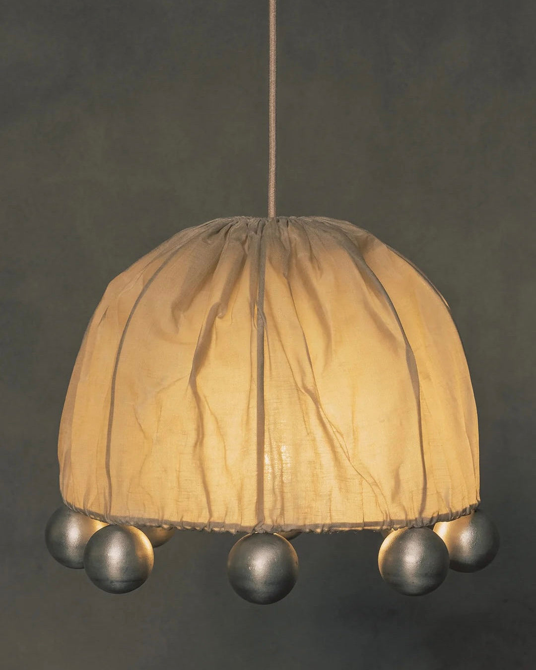 The "ALTER" Pendant Light by Kaytar has a hanging design with a beeswax-coated linen lampshade, featuring a gathered pattern and decorative silver balls around the bottom edge, casting a warm glow against dark backgrounds.