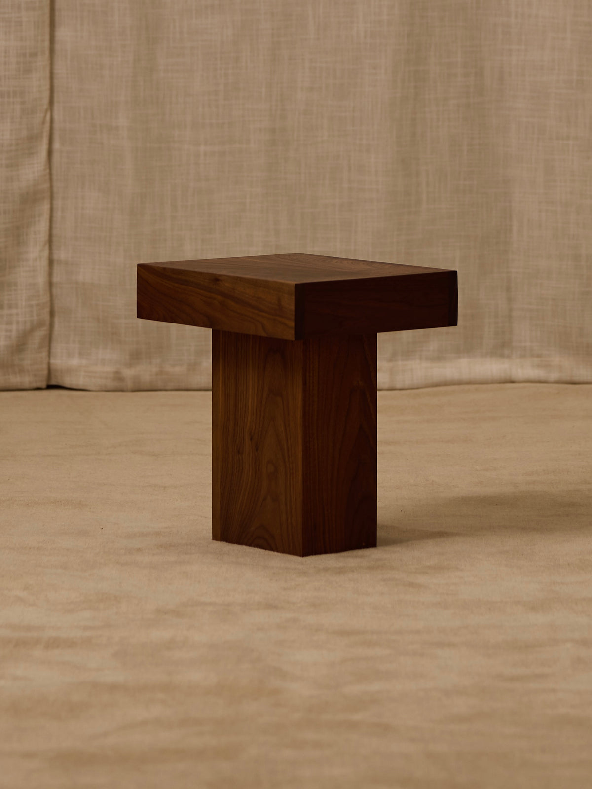 The Jura Pedestal by Ah Um Design Studio, a modern wooden table with a thick square top and solid walnut pedestal base, is displayed on a beige carpet with a textured beige fabric backdrop.