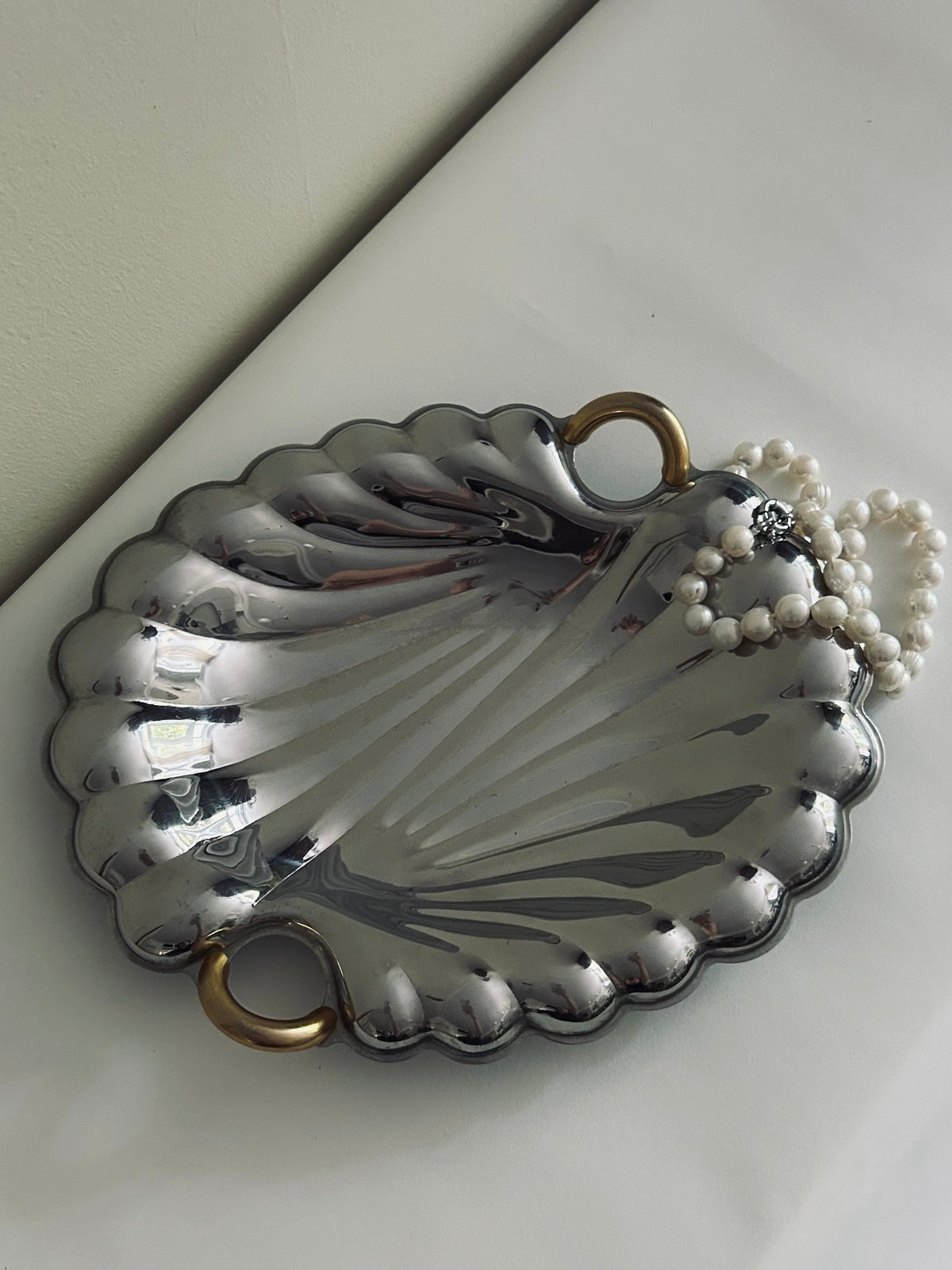 The Italian Stainless Steel Shell Tray by Flock Studio, adorned with golden handles, elegantly cradles a pearl necklace. This luxury design rests on a white surface, reflecting light and casting subtle shadows.