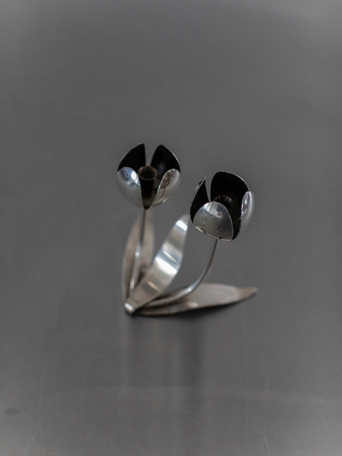 The Steel Tulip Candleholder by Out For Lunch features a sleek steel design that presents a silver metal sculpture of two abstract tulip-like flowers with gracefully curved stems and flat leaves, all set atop a smooth gray surface. This elegant piece contributes a touch of modern home decor, making it ideal for enhancing the aesthetic of any space.