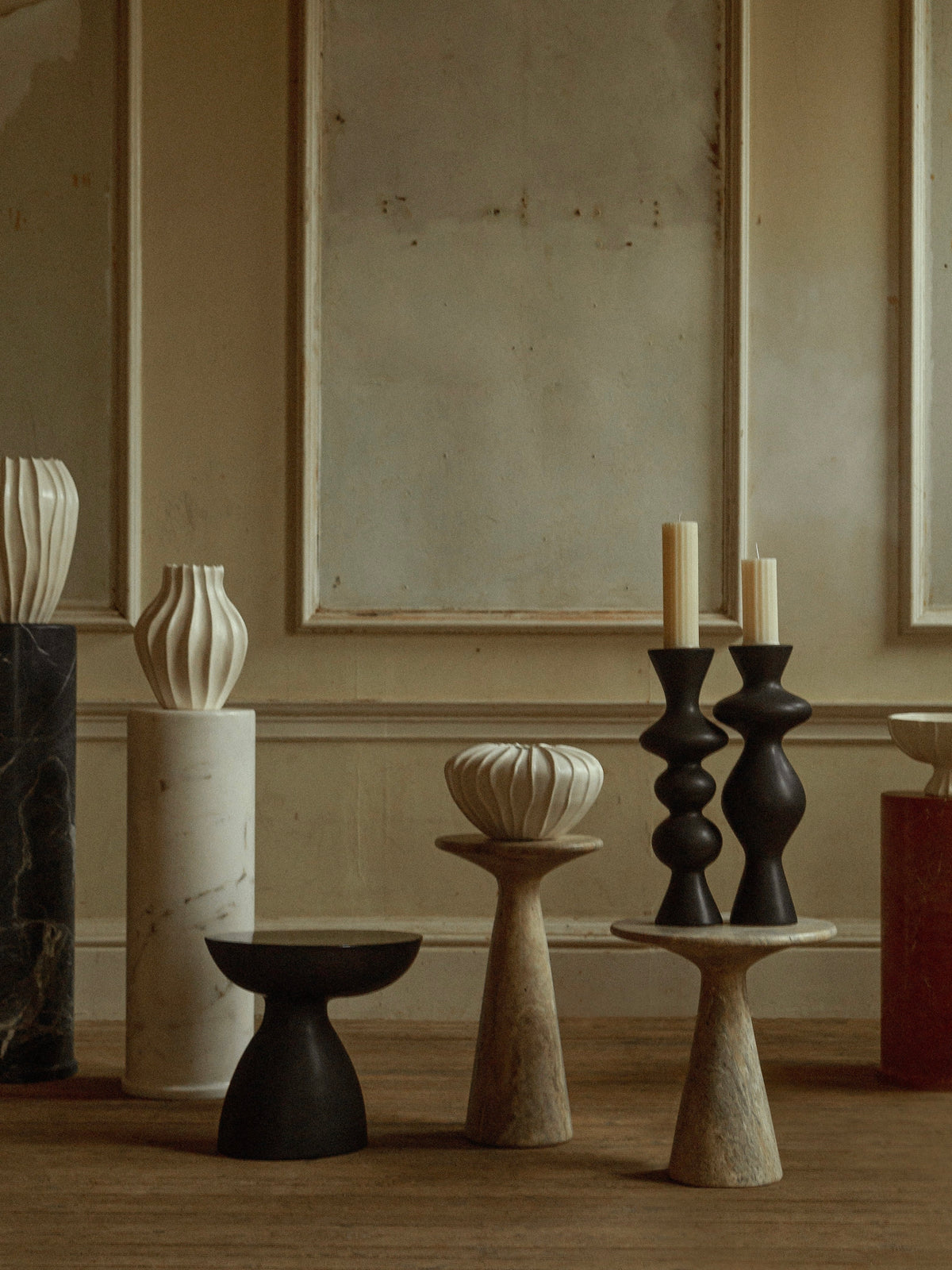 An aesthetic interior scene showcases various stone pedestals and stands, highlighting an asymmetric matte black vase along with candlesticks. The arrangement is beautifully complemented by the "Cleo" Ceramic Side Table from Ransom & Dunn, all set against a textured wall with moldings, while the wooden floor adds warmth to the subdued color palette.