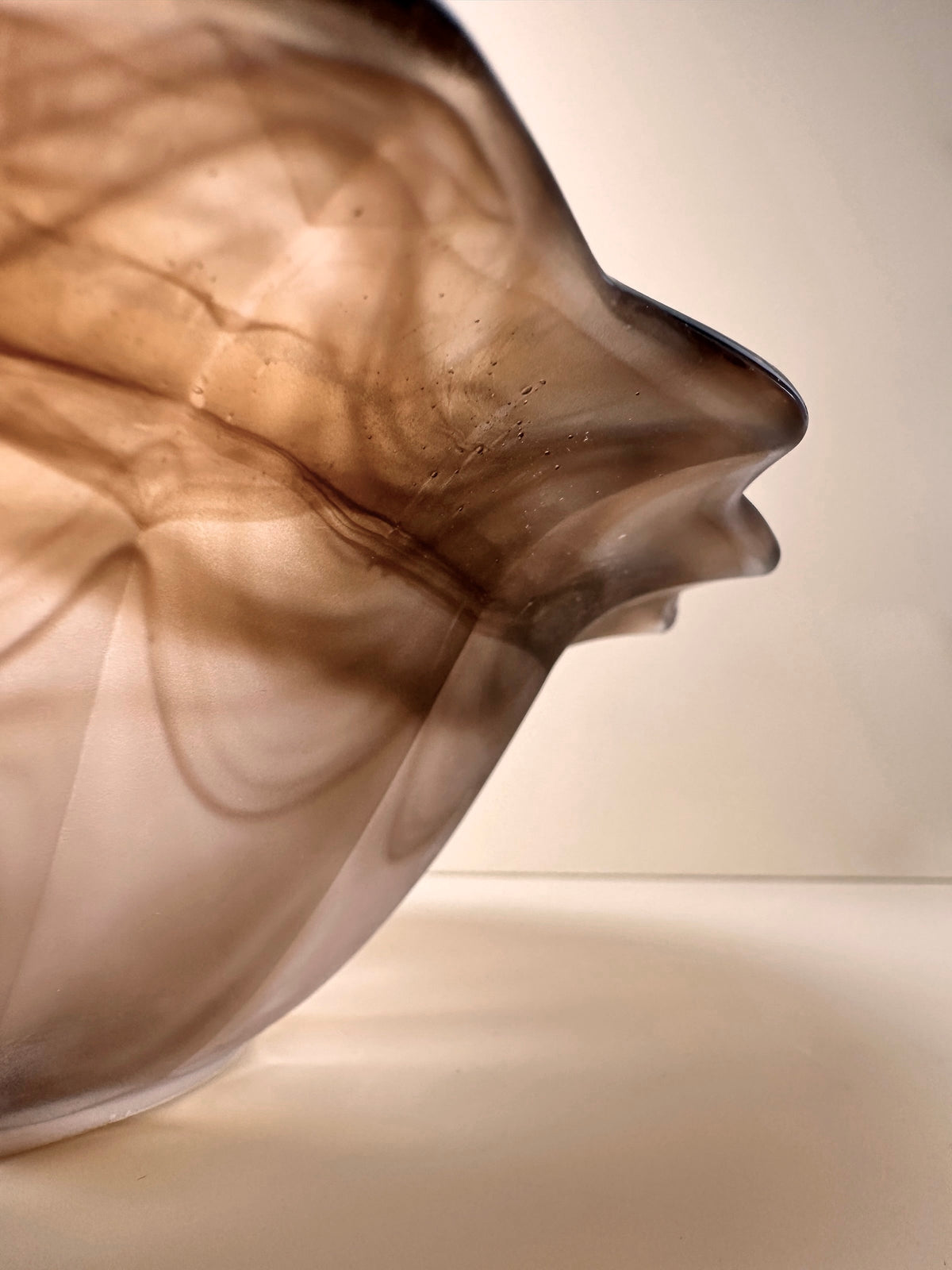 Close-up of a translucent, abstract glass sculpture reminiscent of the German Art Deco Cloud Glass Bowl with pointed rim from around 1930 by Dodo Vintage. The form appears smooth with wave-like patterns and smoky brown and beige tones flowing through it. The plain background emphasizes the intricate details and shapes within the cloud glass.