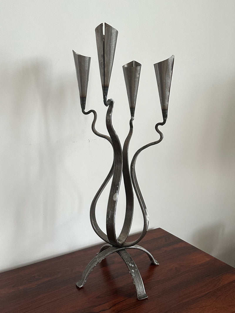The Aesthete Label Post Modern Steel Large Candle Holder features three twisted arms with cone-shaped holders, creating a striking accent for modern decor when displayed on a dark wooden surface against a white wall.