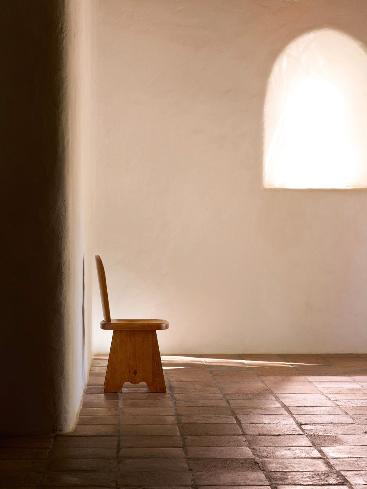 A minimalist room showcases a Lemon's Rambling Chair in Honey French Oak Wood against a plain wall. Soft natural light filters through an arched window, casting shadows on the terracotta-tiled floor, creating an ambiance of simplicity and tranquility that epitomizes contemporary living.