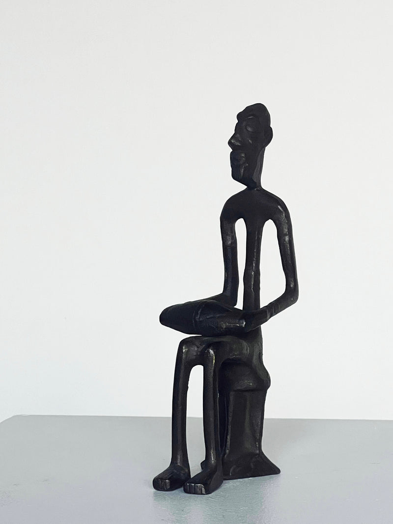 The Bronze Drummer Man Sculpture by Porter Studio is a minimalist black sculpture depicting a seated figure with elongated limbs and an abstract expression, reminiscent of 1980s styles. It resembles a bronze carved man sitting on a stool against a simple white background.