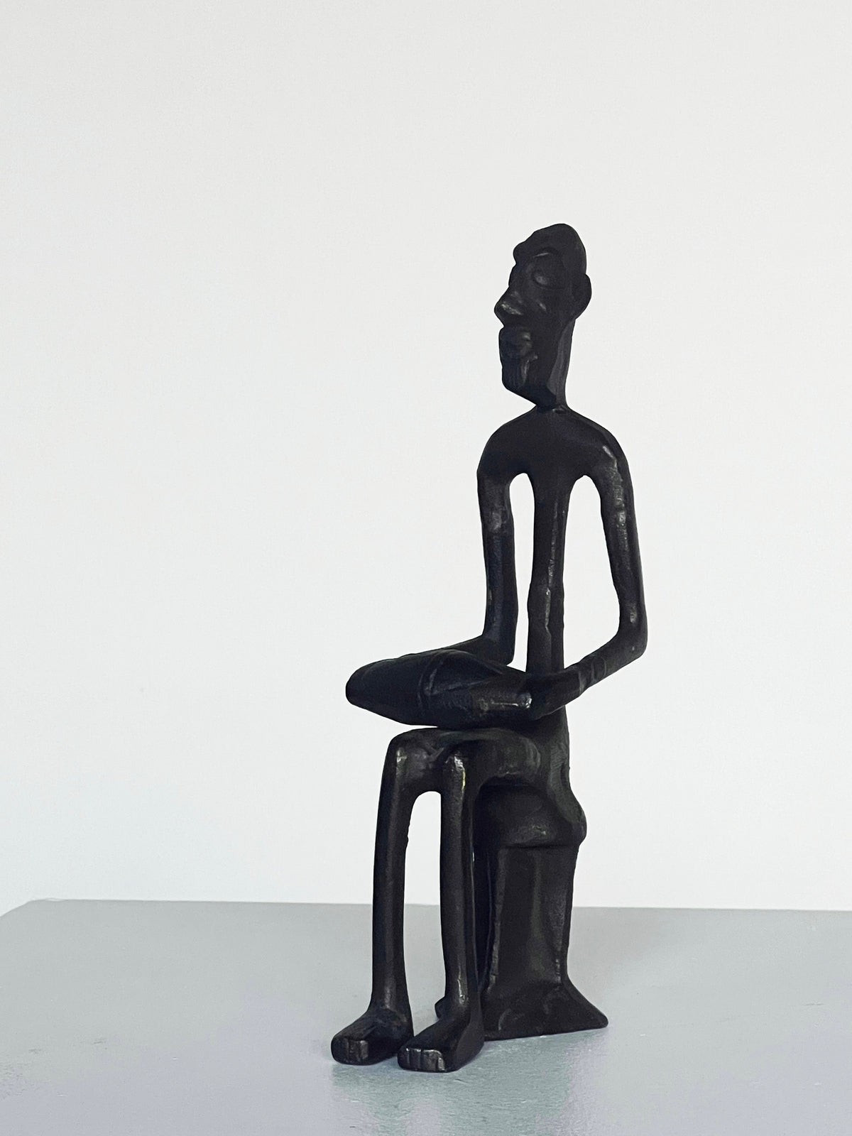 The Bronze Drummer Man Sculpture by Porter Studio is a minimalist black sculpture depicting a seated figure with elongated limbs and an abstract expression, reminiscent of 1980s styles. It resembles a bronze carved man sitting on a stool against a simple white background.
