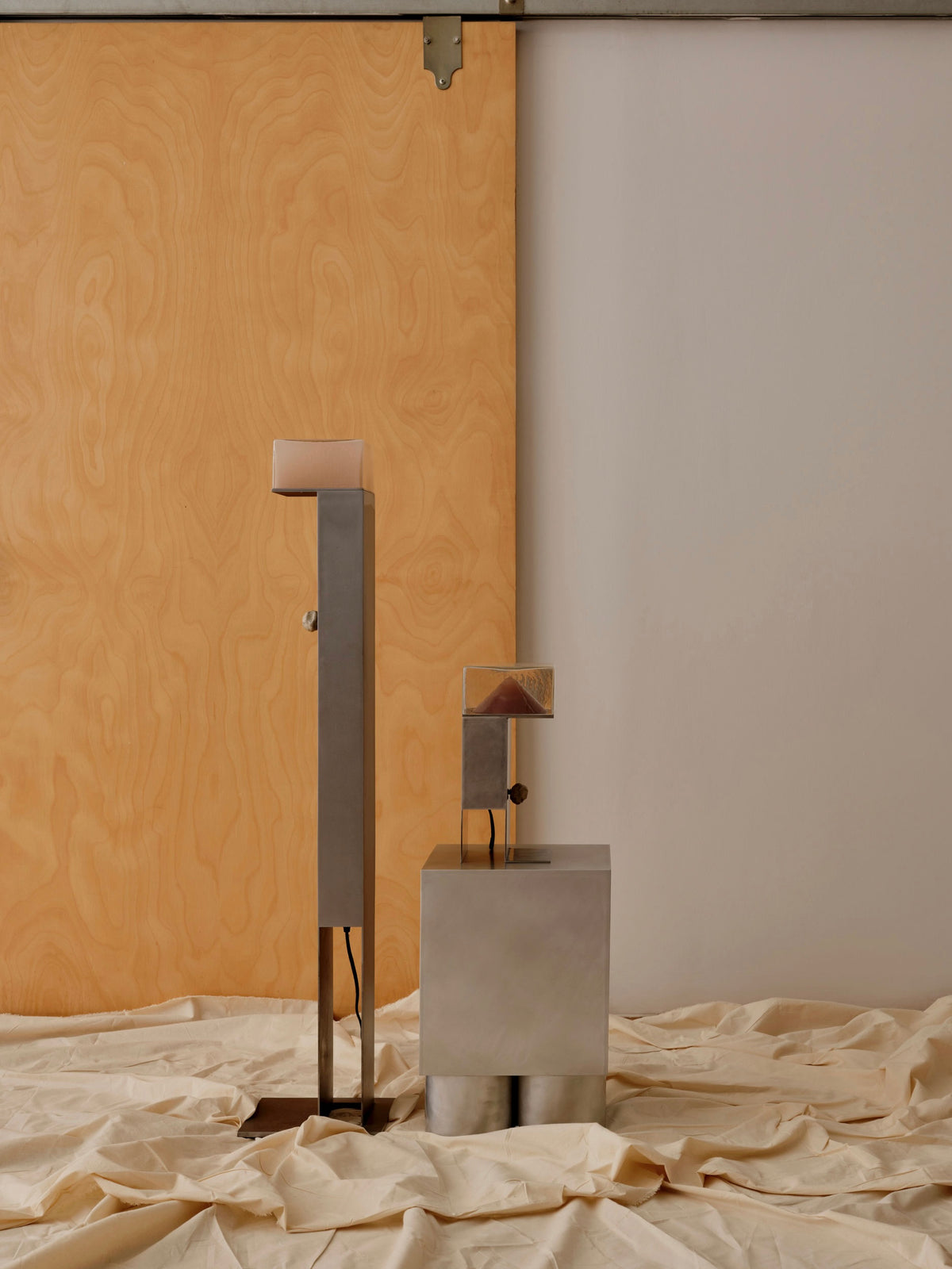 The Perceptions Floor Lamp by Known Work is a minimalist piece featuring two handcrafted abstract metal sculptures with geometric shapes, set on a draped beige fabric against a wooden panel and plain wall for a modern, clean look.
