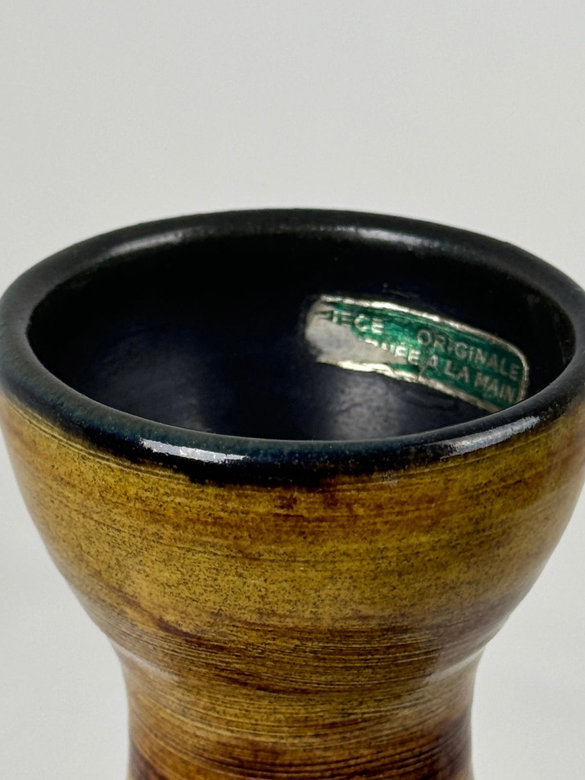 A close-up of a ceramic vase crafted by the potters of Accolay, featuring a glossy dark interior and light brown exterior. Inside, a small label reads "Céramique Originale, Fait à la Main," capturing the charm of incised geometrical decorations often found in rare pieces by Figuresdesign.