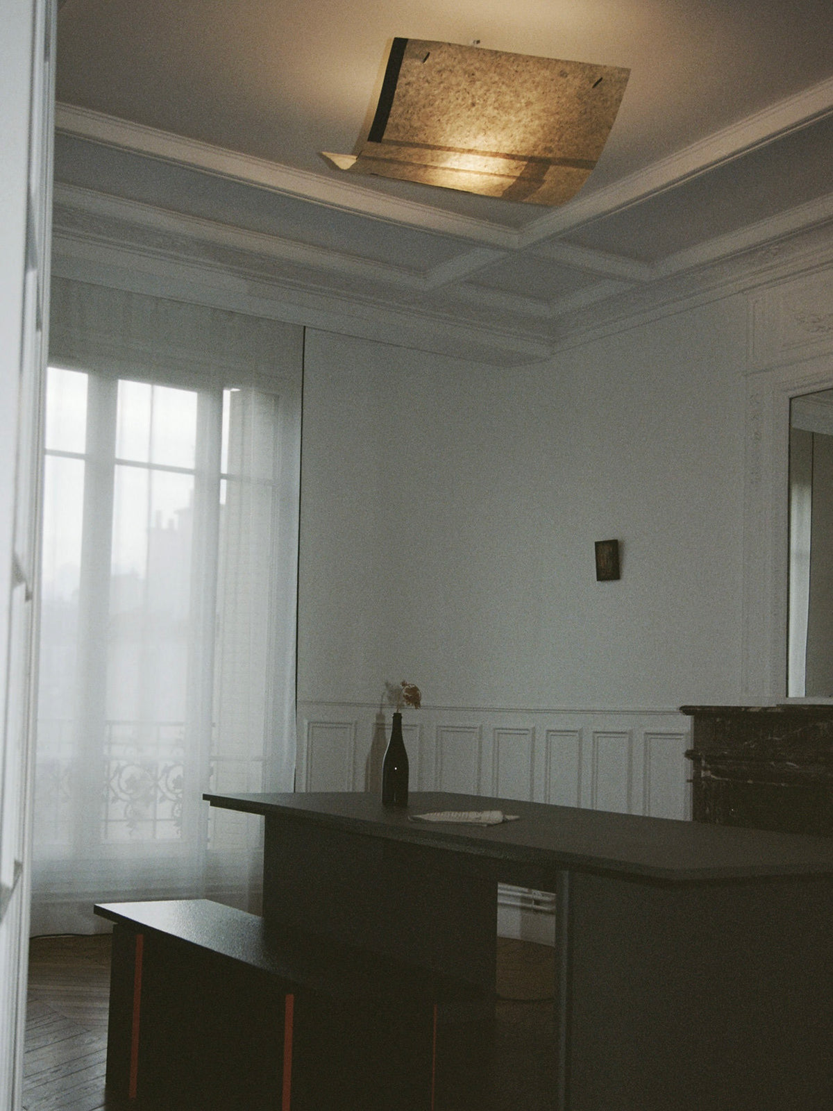 A minimalist room is illuminated by TEGET's handmade Sail Lamp – Rice Field, with soft light filtering through sheer curtains. A dark table holds a wine bottle, glass, and single flower against white walls and parquet flooring. The large window completes the serene setting.