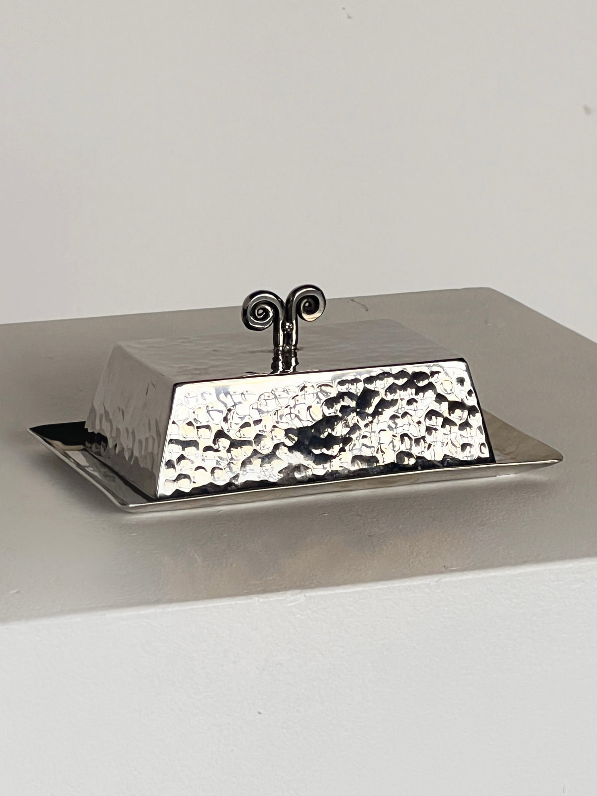 The Spiral Butter Dish by Porter Studio, with a hammered rectangular design and decorative handle, sits on a light surface. The lid exudes 1970s charm, featuring a handle with two outward-facing swirls.