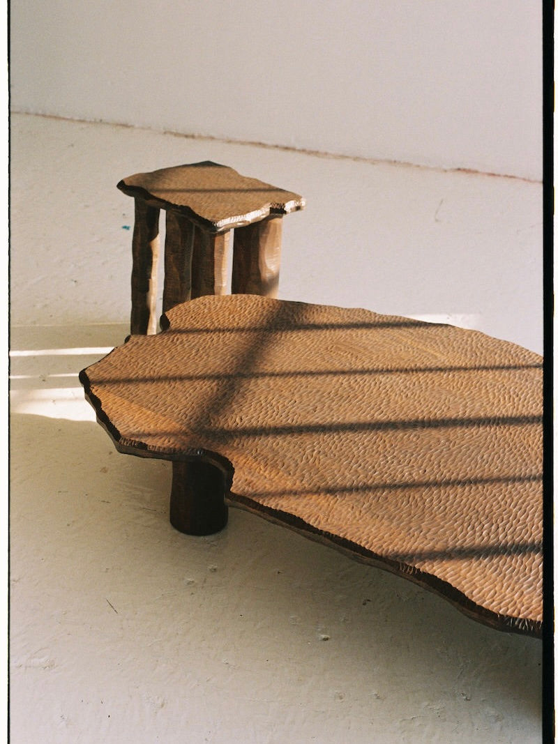 The Mare Coffee Table by Ombia Studio, crafted in hand-carved white oak with organic edges, sits on a light floor as sunlight casts striped shadows across its textured surface.