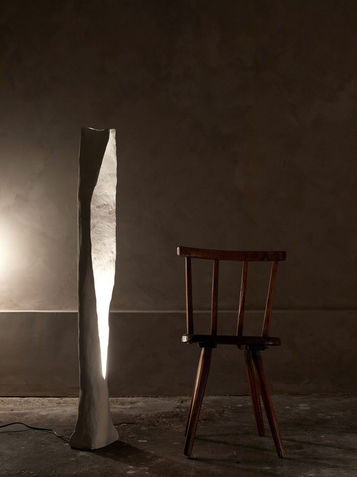 The Alcove Floor Lamp by Studio Tristan Kallas casts a gentle glow over a simple, straight-backed wooden chair, set against a dark textured wall for a minimalistic ambiance.