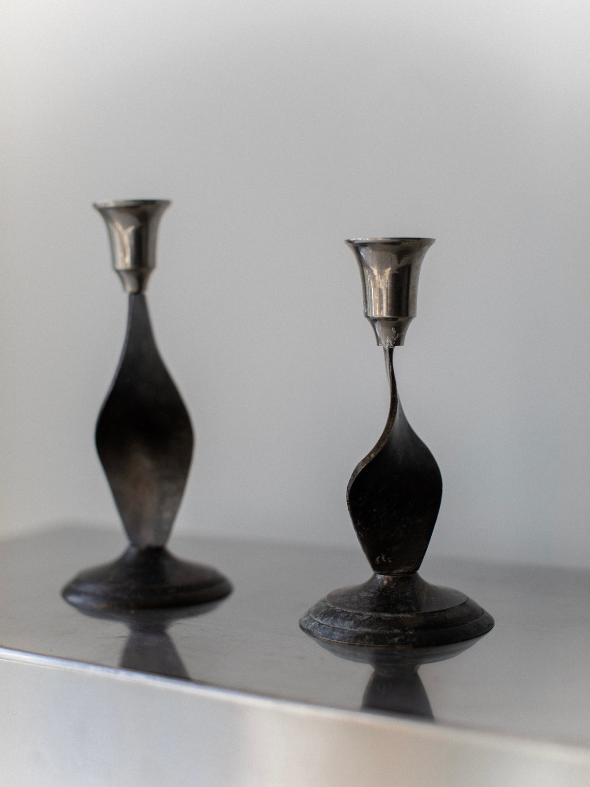 The Brutalist Candleholders by Out For Lunch, made from silver-colored steel with abstract, curved designs, stand elegantly on a reflective surface against a plain background. Inspired by 70's brutalist influences, their simple yet modern aesthetics bring an elegant and minimalist touch to any home decor setting.