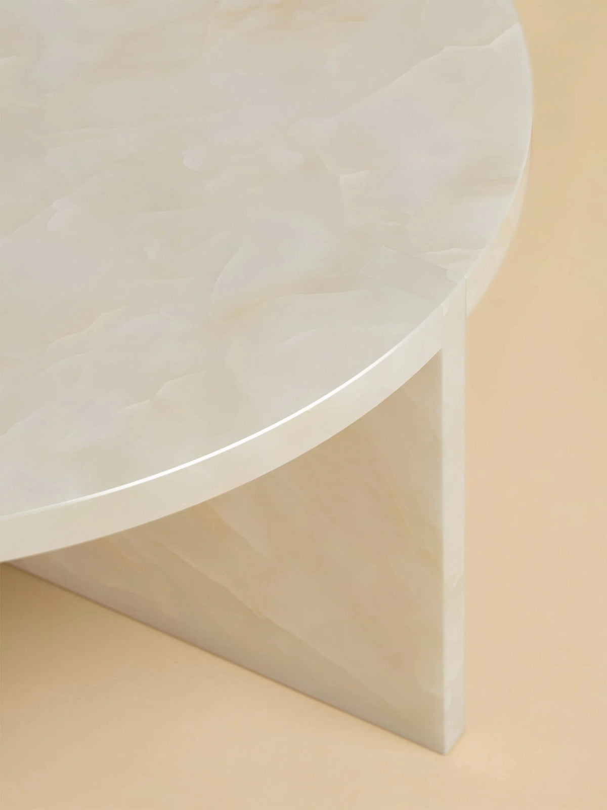 A close-up of the Lebanto "Hashi" Coffee Table reveals a sleek, circular design with a smooth beige marble surface. Its minimalistic look features a single triangular support, blending seamlessly with the soft matching beige background.