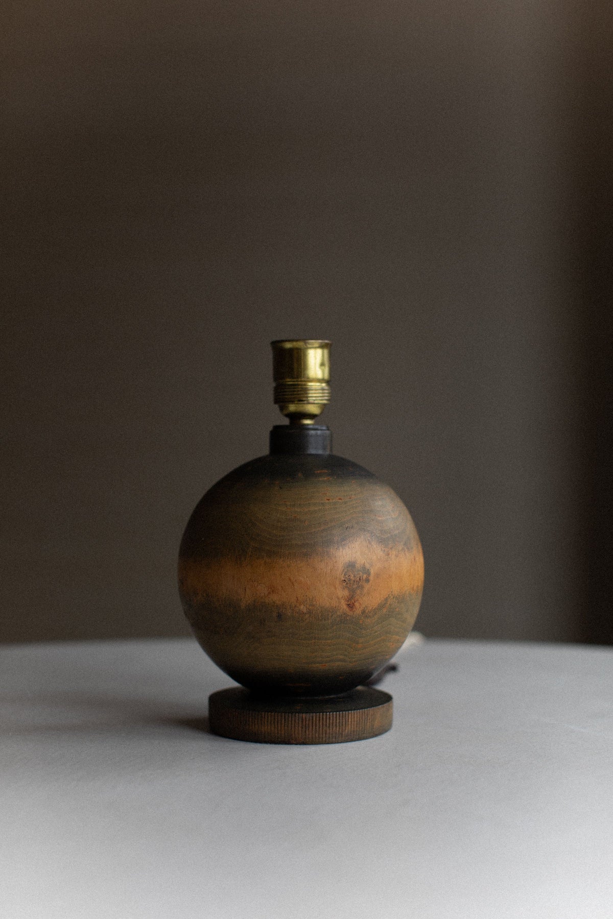 A round wooden table lamp base from the 1920's Globe Lamp by Out For Lunch, featuring a golden socket and no lampshade, sits on a light grey surface. The background is dark and unfocused, accentuating the natural wood grain pattern and exquisite finish of the lamp base. Its timeless design makes it an ideal piece for vintage decor settings.