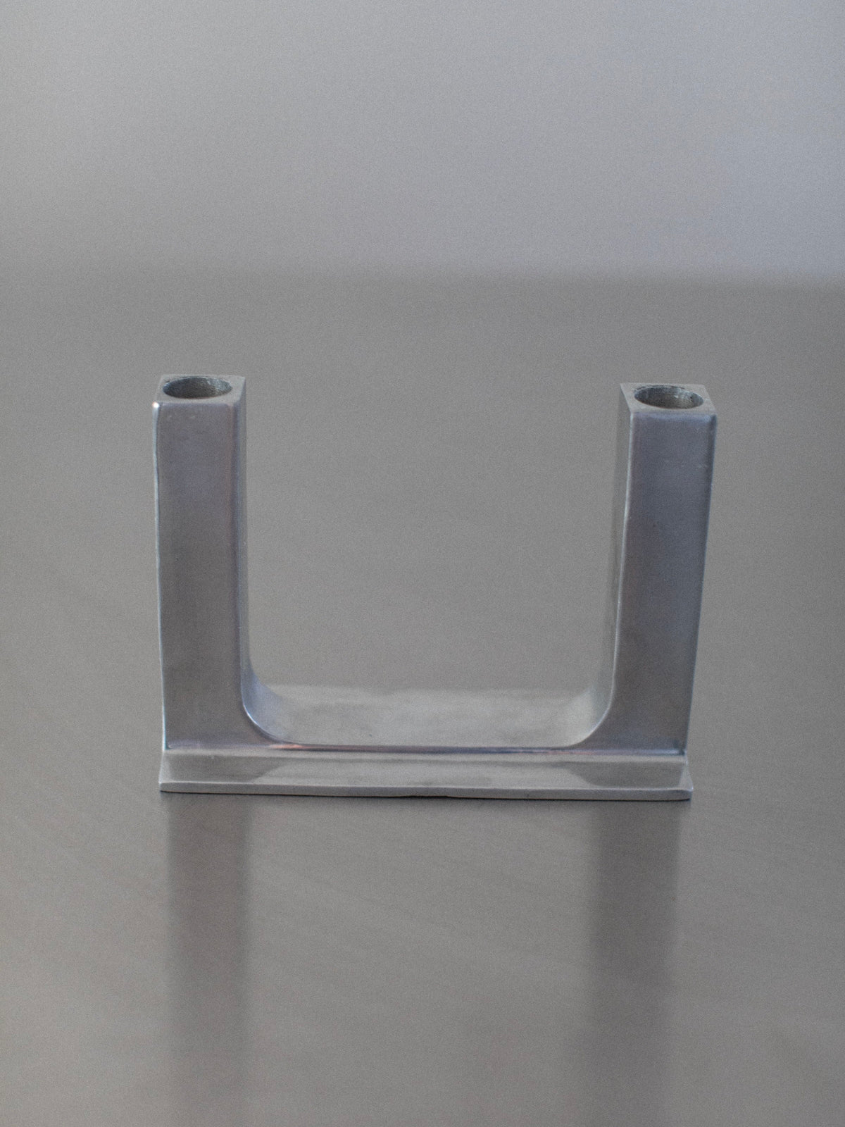 The Chrome Candleholder by Out For Lunch showcases a striking U-shaped design with two vertical cylindrical sections at each end, resting elegantly on a smooth, reflective surface. The backdrop is subtly blurred, casting a neutral, light grey hue that enhances its brutalistic elegance.