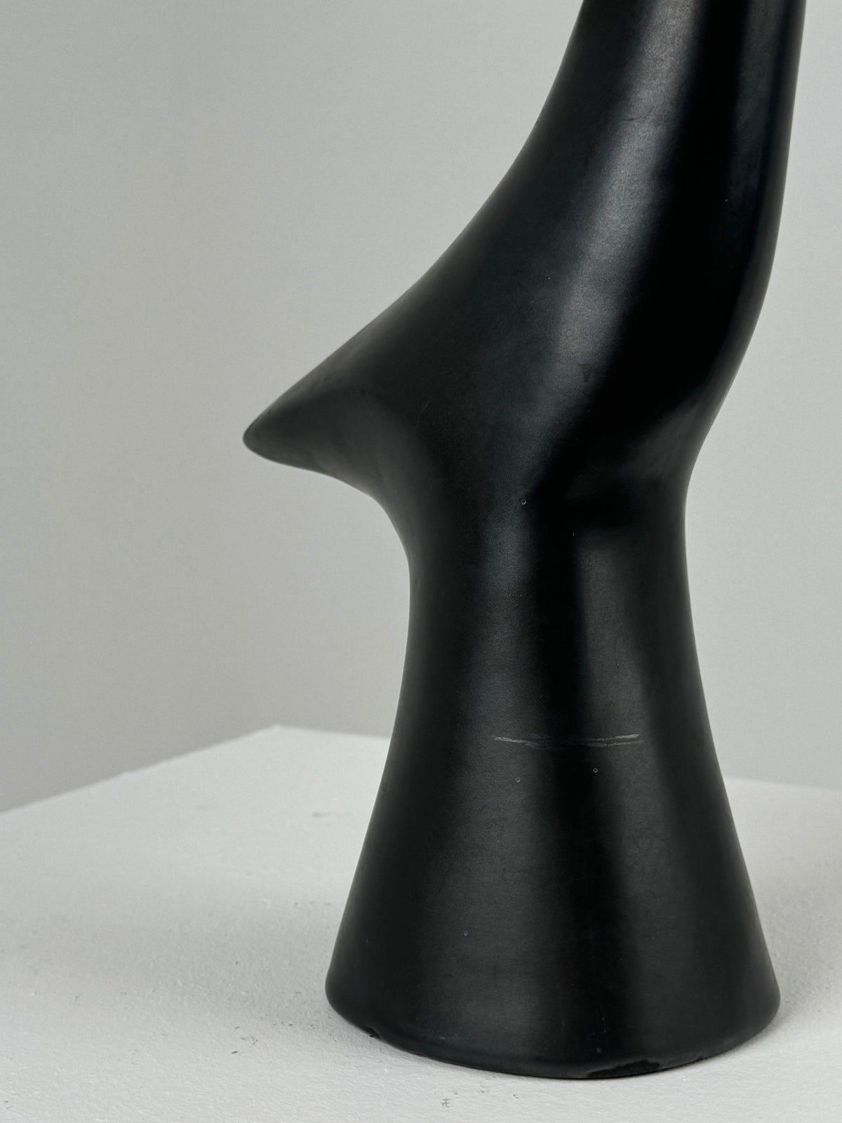 Displayed on a white pedestal against a plain background is the sleek Zoomorphic Ceramic Lamp by Figuresdesign. This black glazed ceramic piece features a smooth, curved form reminiscent of simplified, elongated bird or swooping figure designs from France in the 1960s.