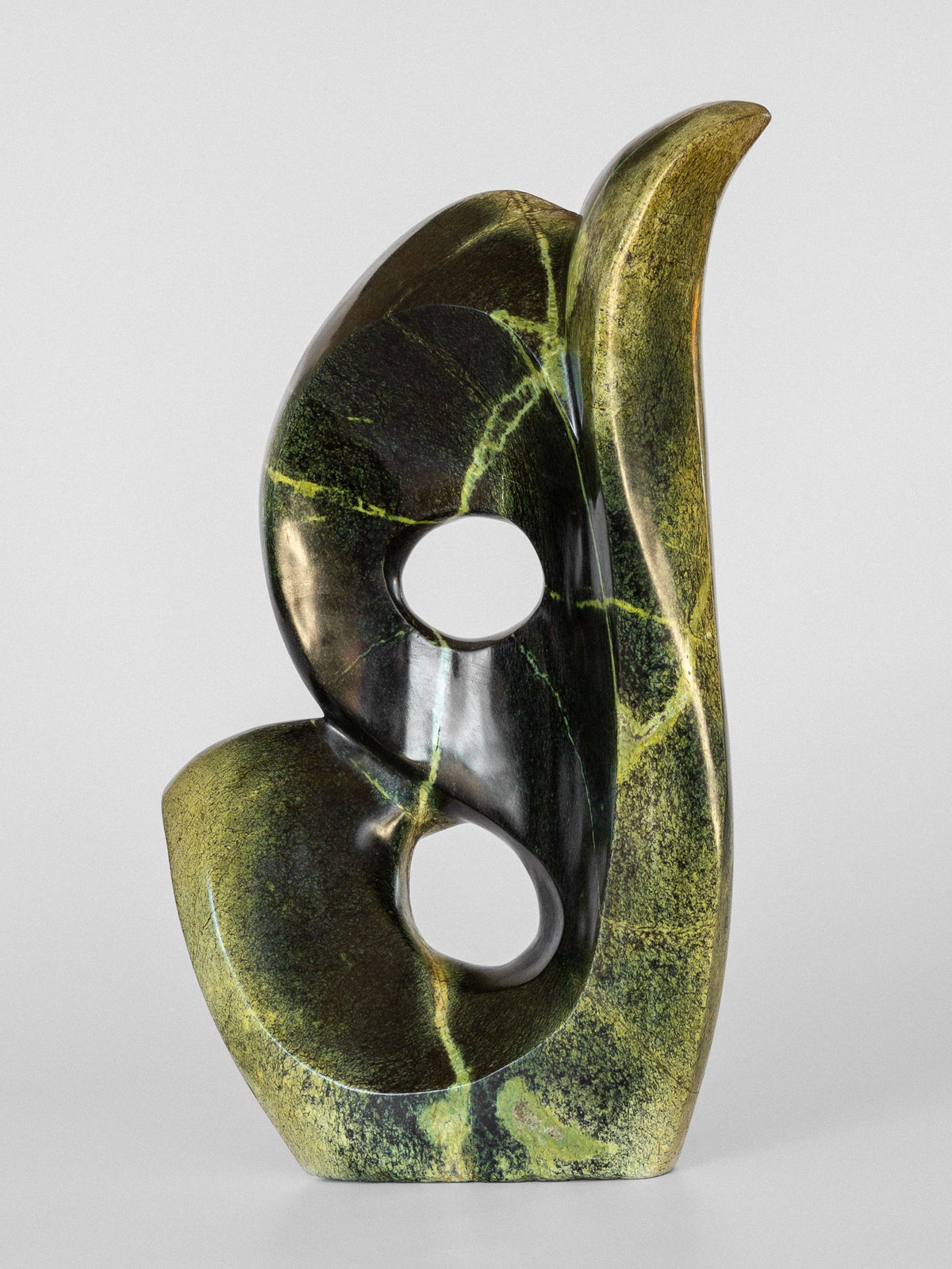 Abstract Stone Sculpture II by Spigel features smooth curving forms with two circular openings resembling intertwined shapes. This Zimbabwean-crafted piece in green and black has a polished leopard stone surface that highlights its texture and color variations.