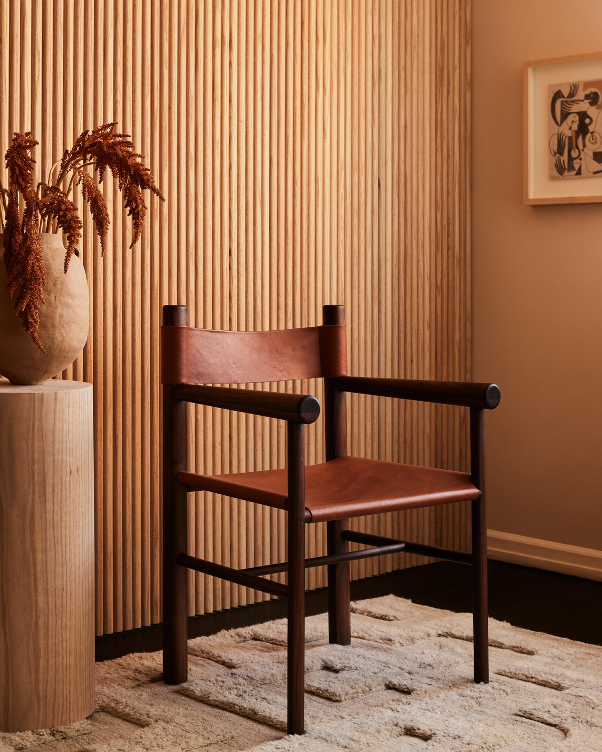 A minimalist room features a brown leather chair with wooden arms on a textured cream rug. A marta bonilla Column Vase, crafted in terracotta stoneware with dried plants, rests on a wooden pedestal. Vertical wooden slats and framed abstract art complete the background.
