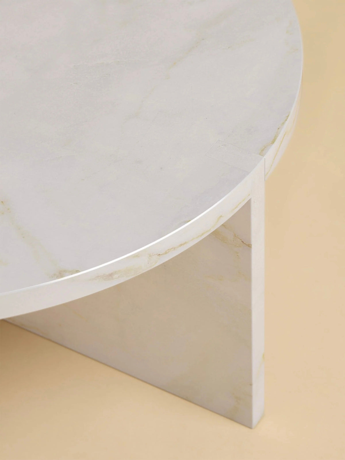Close-up of the "Hashi" Coffee Table in Marble by Lebanto, featuring a circular top with subtle gray veining on its white surface and a triangular base. It's set against a neutral beige background.