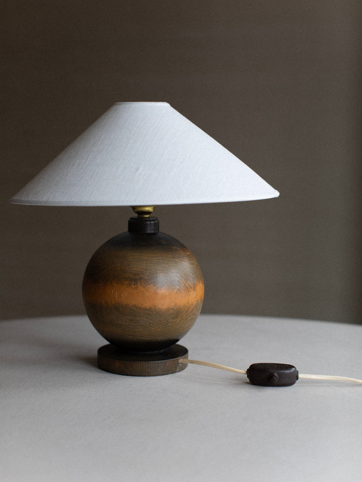 The 1920's Globe Lamp by Out For Lunch, featuring a round wood finish base and a conical white lampshade, sits on a light-colored surface. The lamp's cord and switch are visible, resting on the table next to it, enhancing its vintage decor charm. The background is blurred and neutral in color.