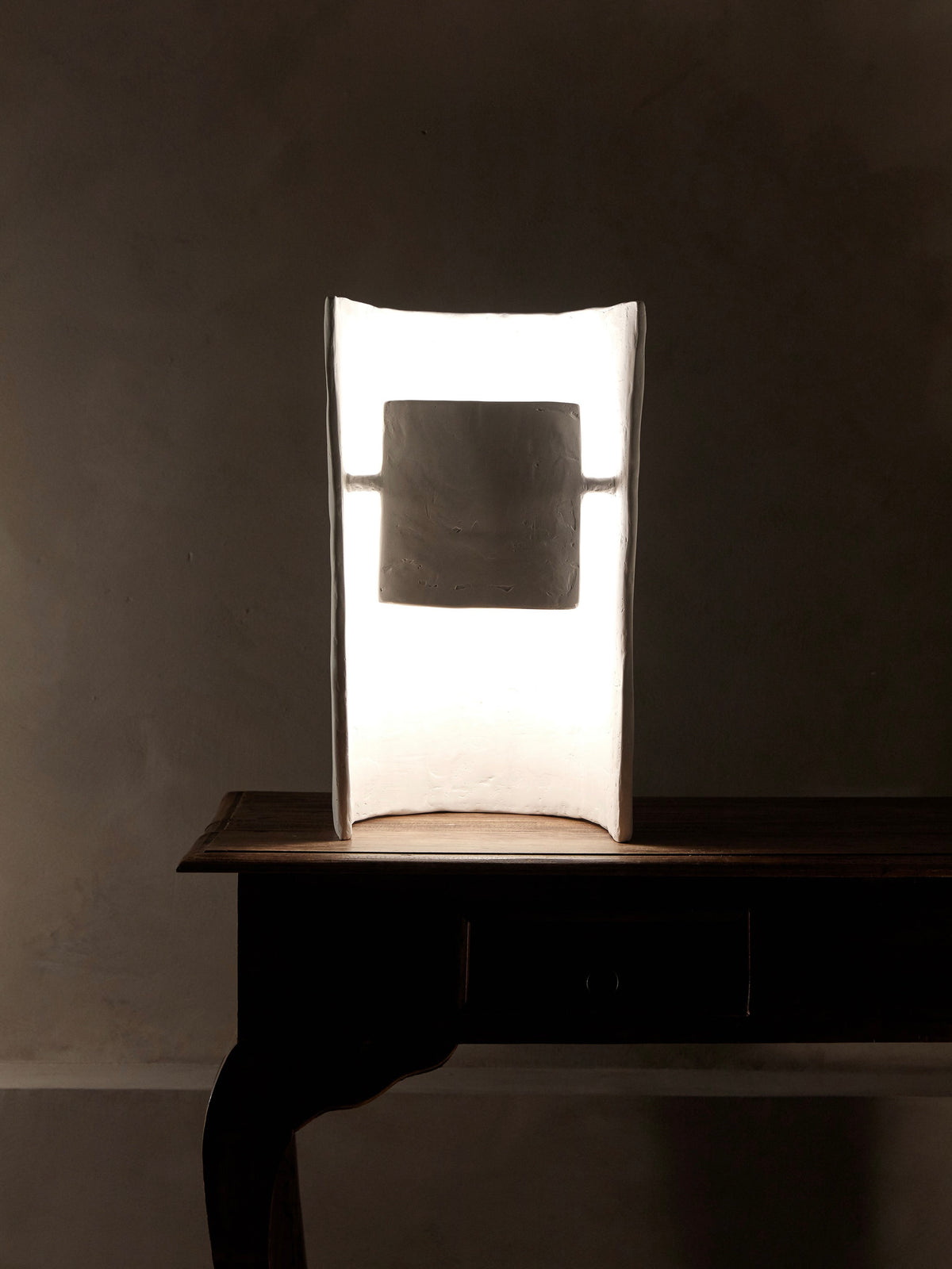 The Crescent Table Lamp by Studio Tristan Kallas, featuring a minimalist design and a thin rectangular shade, rests elegantly on a wooden table against a dark backdrop. Its soft white light offers indirect illumination, creating an ambient setting reminiscent of fashion photography.