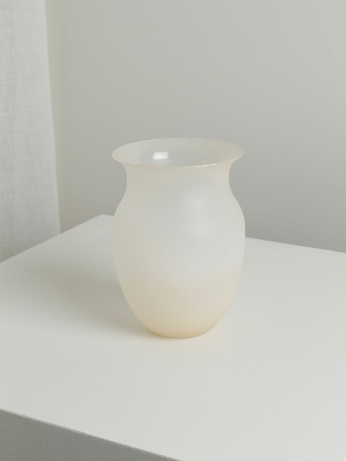 A medium vase in Sahara from Verre d'Onge, crafted with a minimalistic design out of mouth-blown frosted white glass and featuring a slightly flared rim, sits centered on a plain white pedestal against a Sahara-toned background. The scene exudes a clean, modern aesthetic with soft, diffused lighting.
