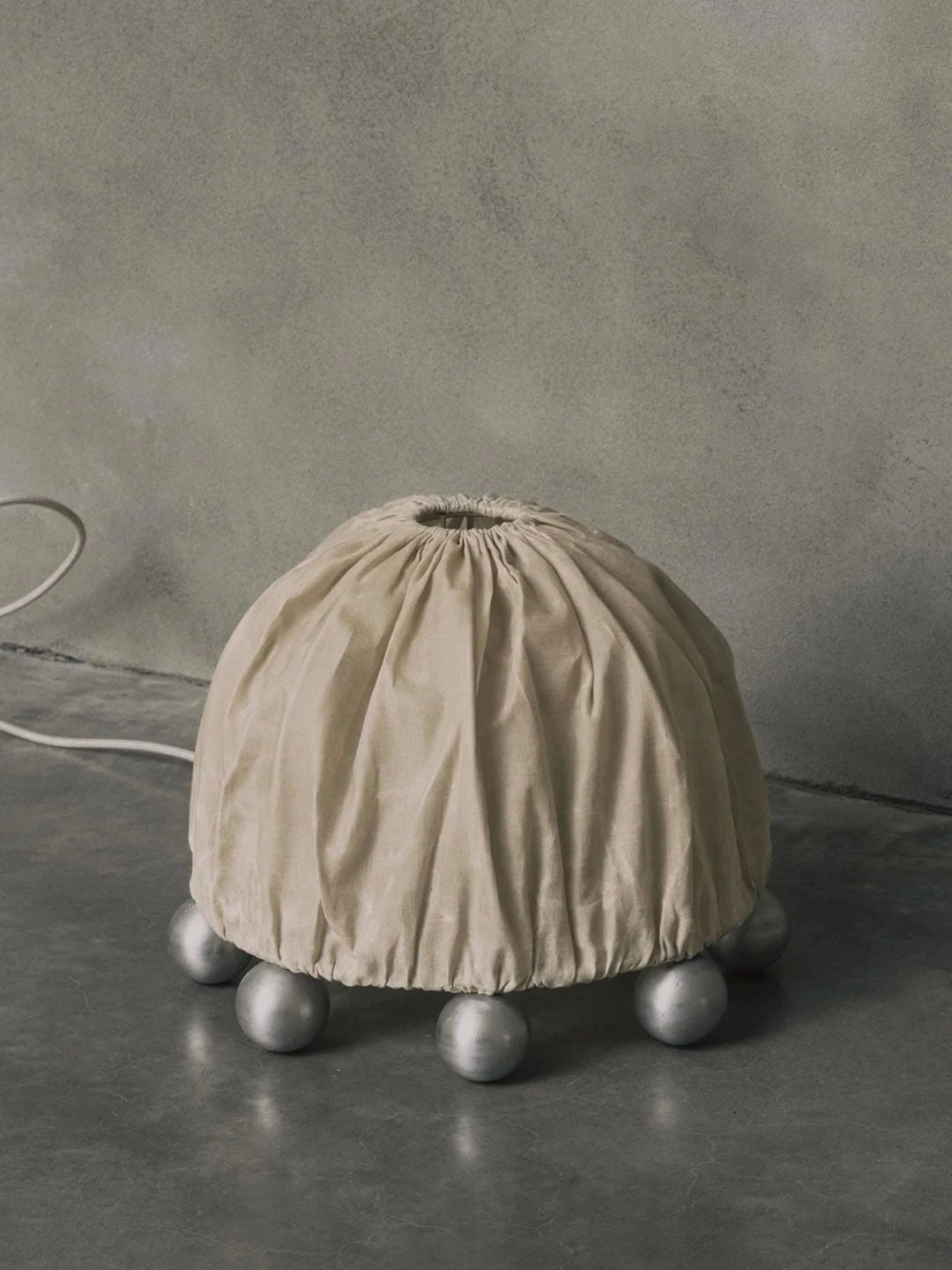 The Kaytar "ALTER" Table Lamp features a beige linen dome top, cinched above six metallic spheres. Set against a textured gray wall, the cord extends across a smooth gray surface, fusing balance and design in this sculptural piece made of aluminium and beeswax.