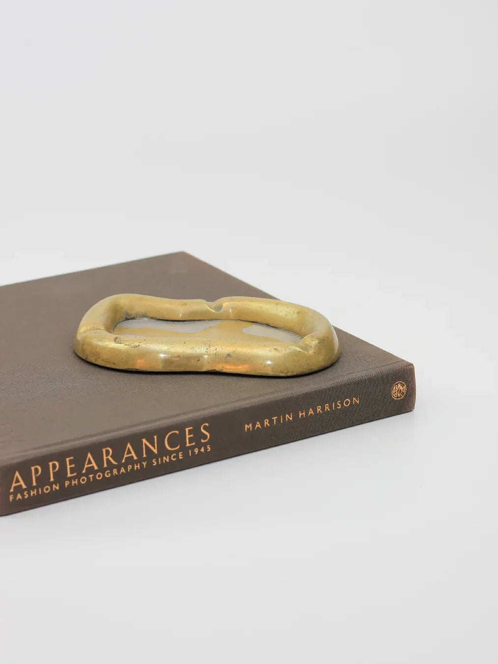 The book "Appearances: Fashion Photography Since 1945" by Martin Harrison rests on a white surface, accompanied by the Brutalist Ash Tray from Boga Avante Shop, featuring a rustic brass finish in an abstract shape.