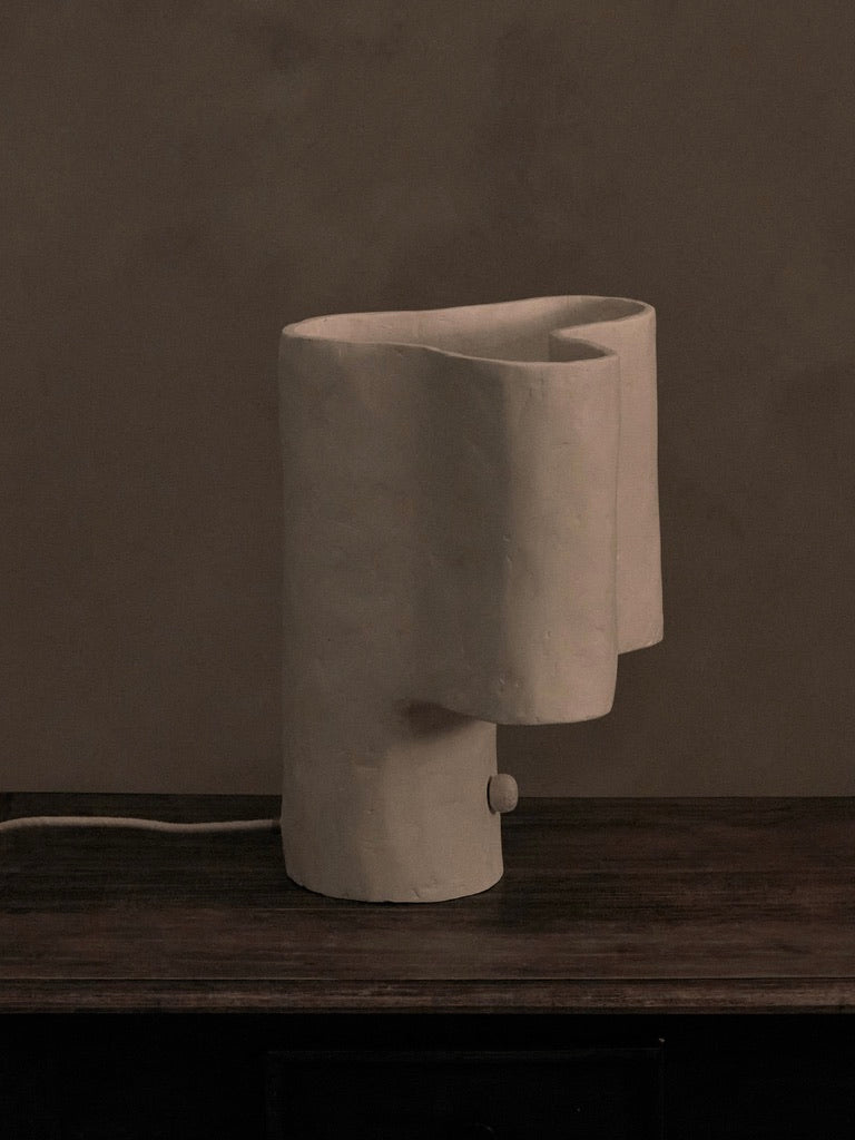 The Cantilever Freeform Table Lamp by Studio Tristan Kallas is a minimalist, sculptural piece crafted from smooth, white material with a design resembling draped fabric. Handcrafted with precision, it elegantly complements a dark wooden surface against a neutral background.