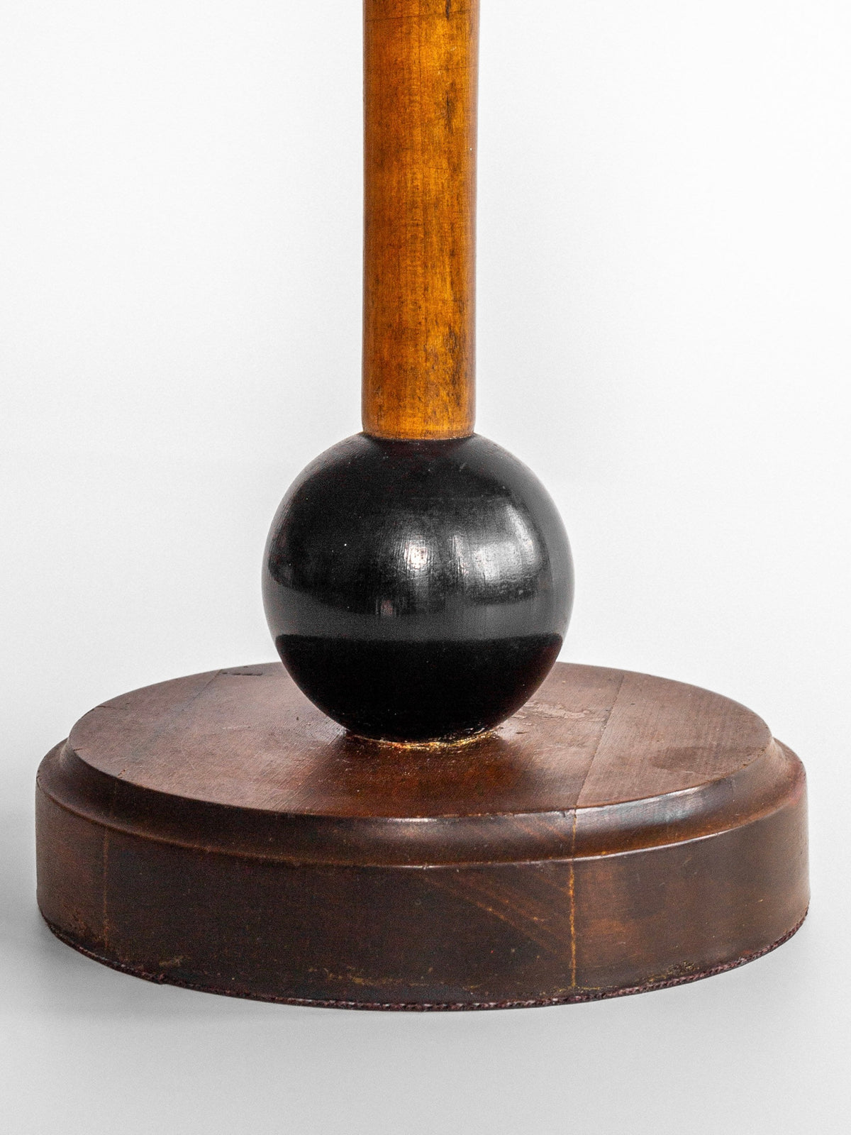 A close-up photograph showcases a Spigel Wooden Art Deco Table Lamp, inspired by 1930s Swedish design. The base is circular and made of dark wood, with a centrally attached black-painted sphere. Extending upward from the sphere is a wooden rod that captures the elegance of Art Deco table lamps. The background is plain and gray.