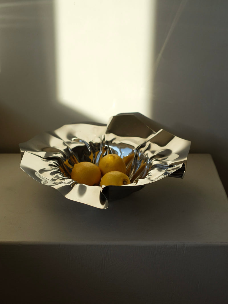 The Folded Chrome Bowl by septembre studios, with its shiny metallic finish and unique folded shape, elegantly holds three lemons. Placed on a white surface, the bowl is beautifully illuminated by soft natural light, creating delicate shadows on the wall behind—making it a perfect accent for any interior space.