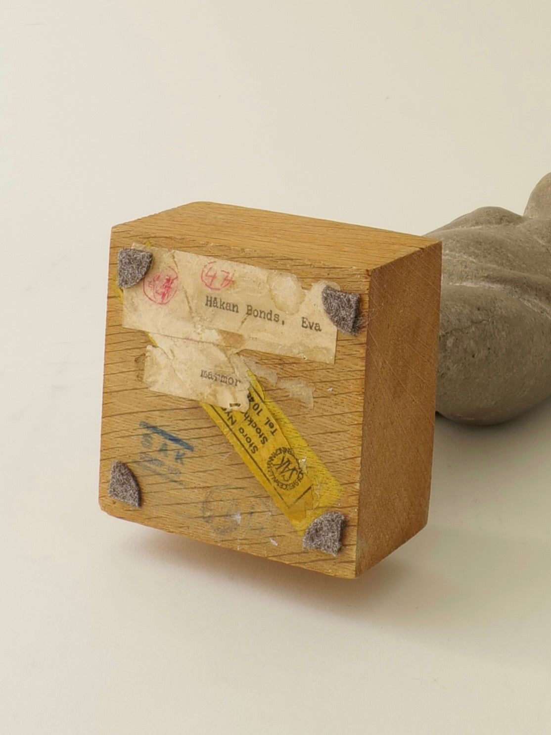 A wooden block with worn labels and corner protectors sits on a white surface, suggesting ties to the "Marble Figurine Sculpture by Håkan Bonds" from Collection apart, with part of a gray textured object visible behind it.