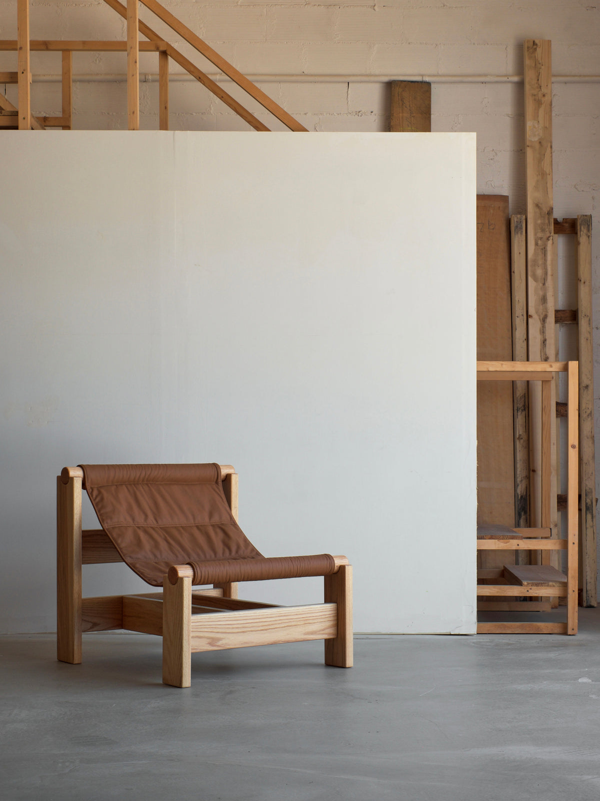 The malva "Lounge Up" Lounge Chair features a wooden frame and brown leather seat, delivering adaptive comfort in a minimalist space with unfinished wooden beams, white walls, and a concrete floor.