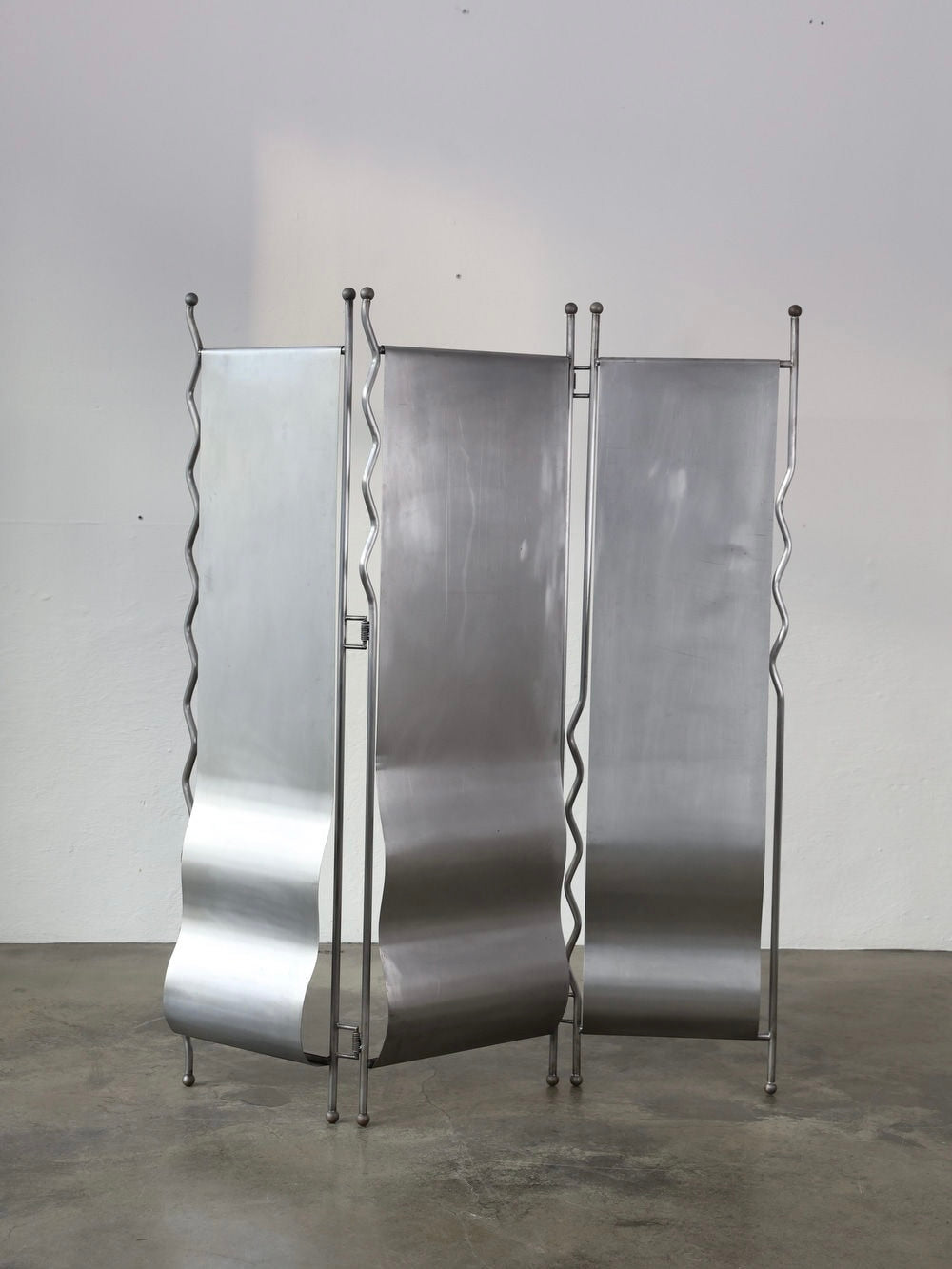 The sleek and contemporary "Alto" folding screen by The Good Living&Co., made in Poland from shiny stainless steel with a wavy design, stands on a gray concrete floor against a plain white wall, reflecting light beautifully.