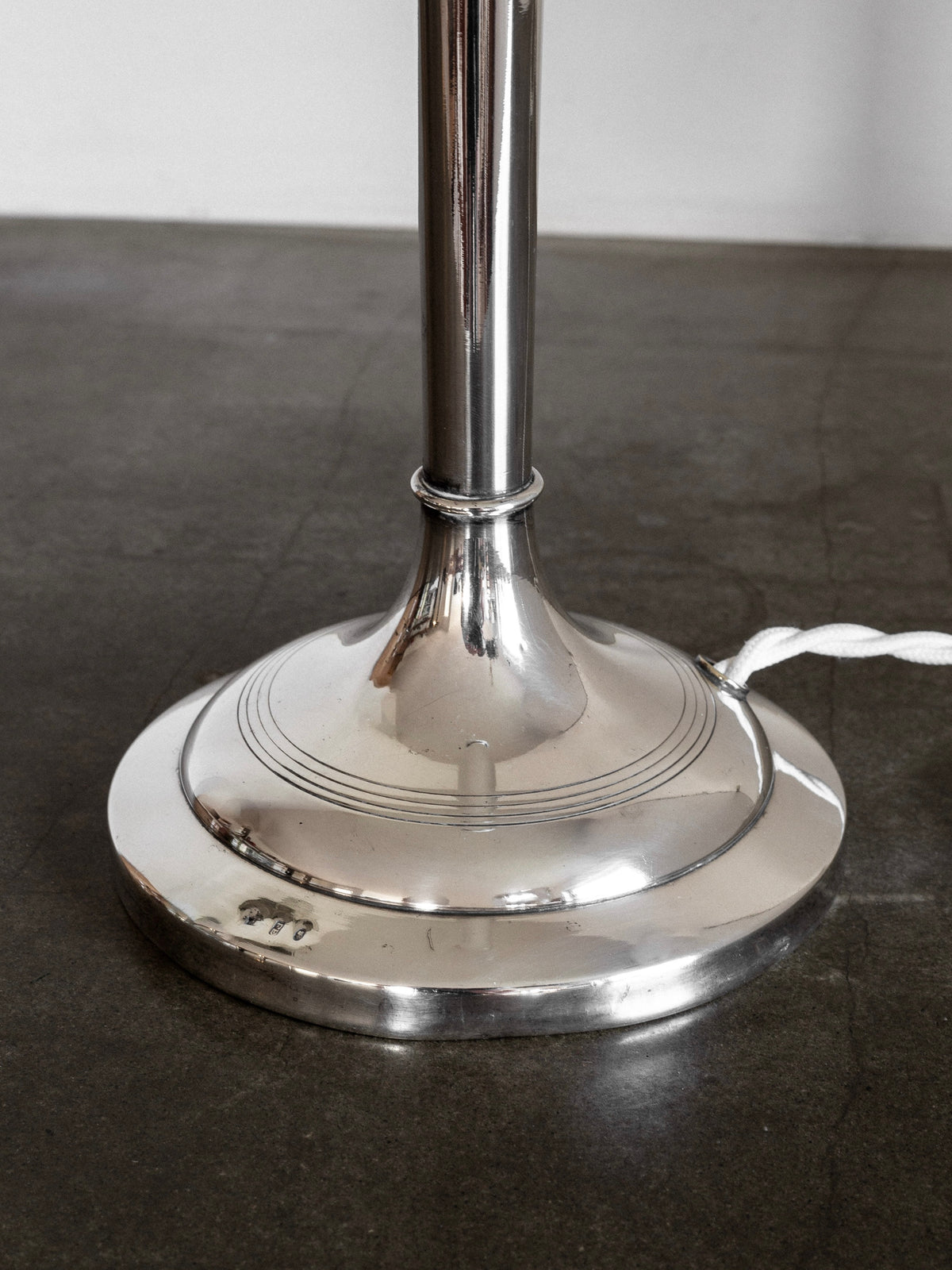 A close-up view of the metallic base of the Silver-Plated Table Lamp, Early 1900s by Spigel, standing on a dark floor with neoclassical influences. A white electrical cord extends from the right side of the lamp base. The reflective surface shows signs of wear, adding character to its polished look.
