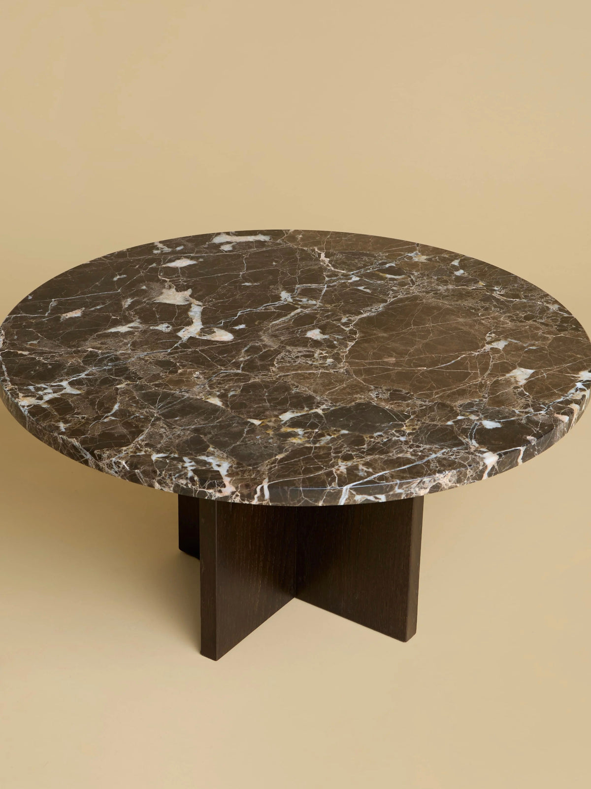 The "Tinian" Coffee Table by Lebanto features a round, dark marbled stone top and a sleek cross-shaped base made from rich oak wood, set against a plain beige background.