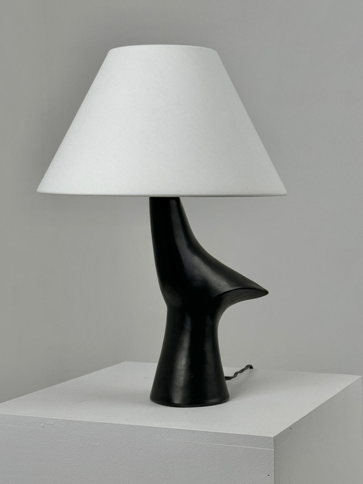 The Zoomorphic Ceramic Lamp by Figuresdesign features a sleek black base that resembles an abstract sculpture, topped with a minimalist white conical lampshade. Made from black glazed ceramic, it is displayed on a white pedestal against a plain gray background, capturing the sophisticated design ethos of 1960s France.