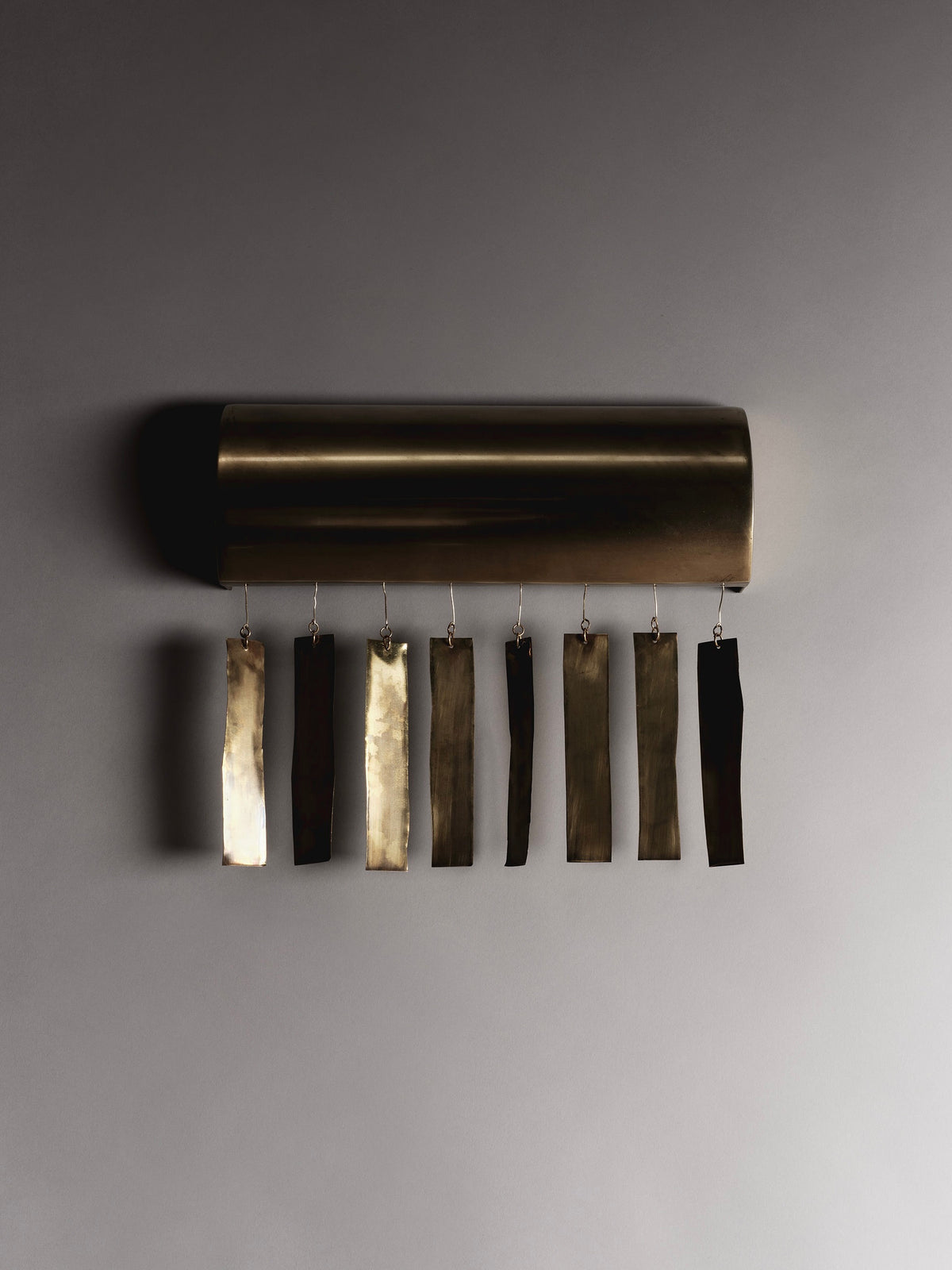 The Ombia Studio Quimbaya Sconce is a handmade brass wall fixture featuring eight dangling metal rectangles in gold and bronze shades. Mounted on a smooth gray wall, it casts gentle shadows with its cylindrical base design.