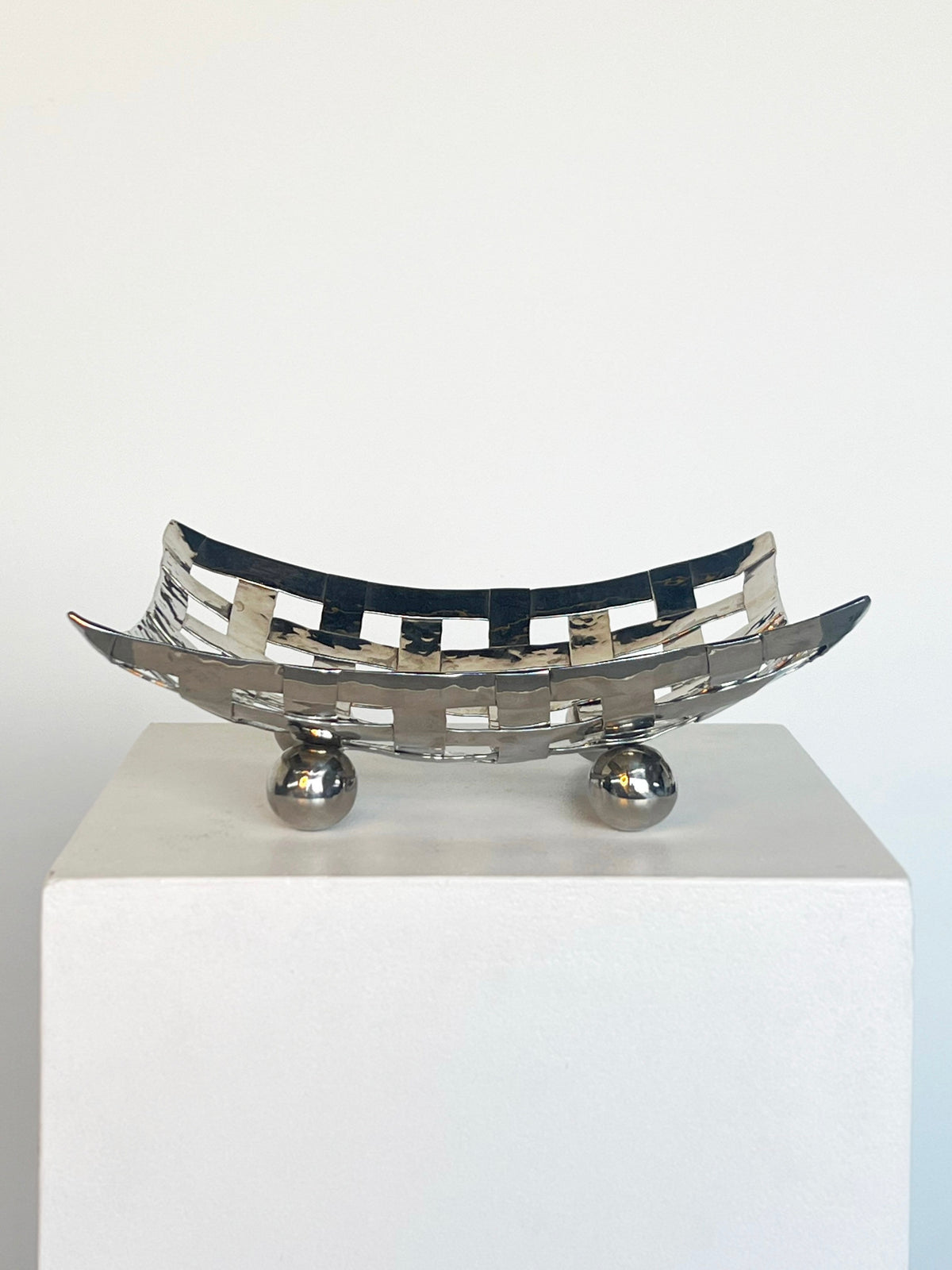 The Porter Studio Aluminum Dish on Feet, with a woven pattern and two ball feet, is showcased on a white pedestal. Its reflective silver finish contrasts elegantly against the plain background, highlighting its beautiful proportions.