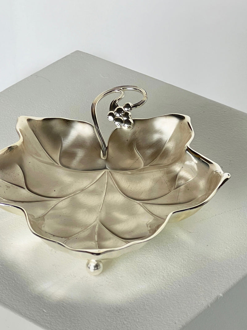 The Porter Studio Grape Leaf Dish, a vintage 1930s silver creation, resembles a grape leaf vine tray with its curled stem handle. It elegantly perches on small ball feet atop a square pedestal, while the intricate vein details and polished surface beautifully capture and reflect light.