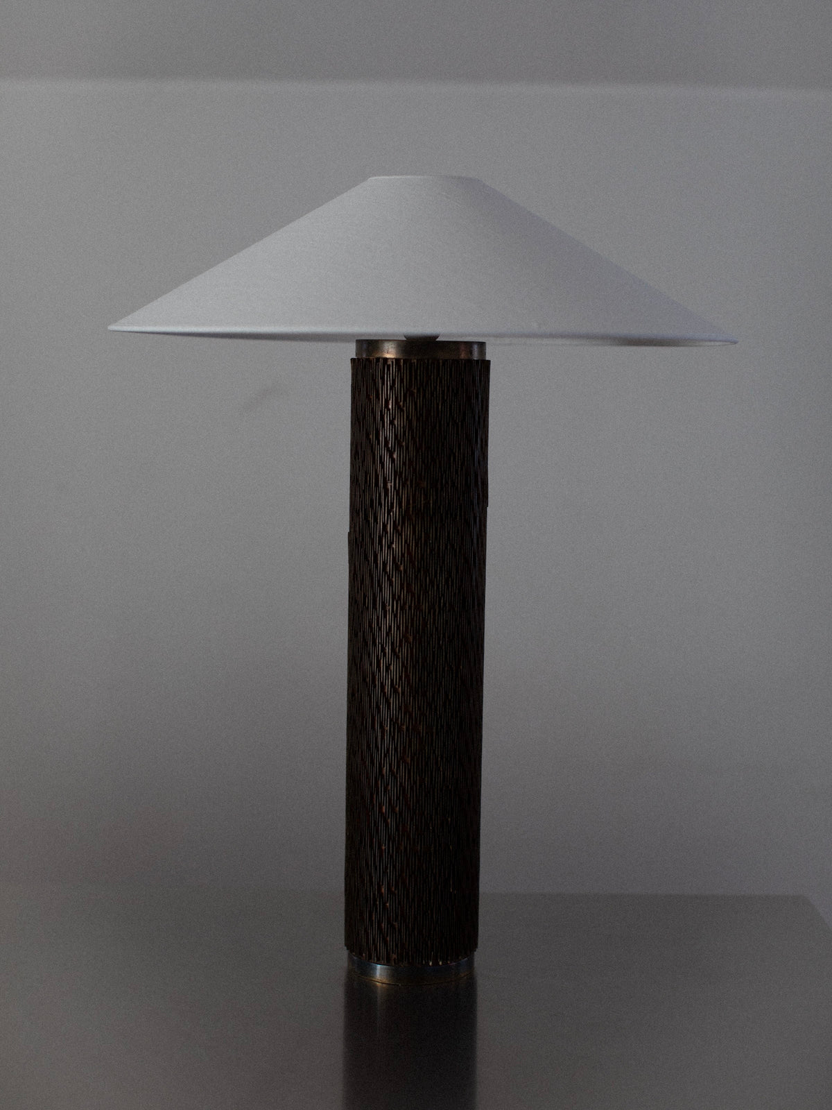A modern Brutalist Floor Lamp by Out For Lunch, featuring a cylindrical dark-textured base and a wide conical white lampshade, is positioned on a metallic surface against a plain grey background. The lamp combines contemporary design elements with a minimalist aesthetic.