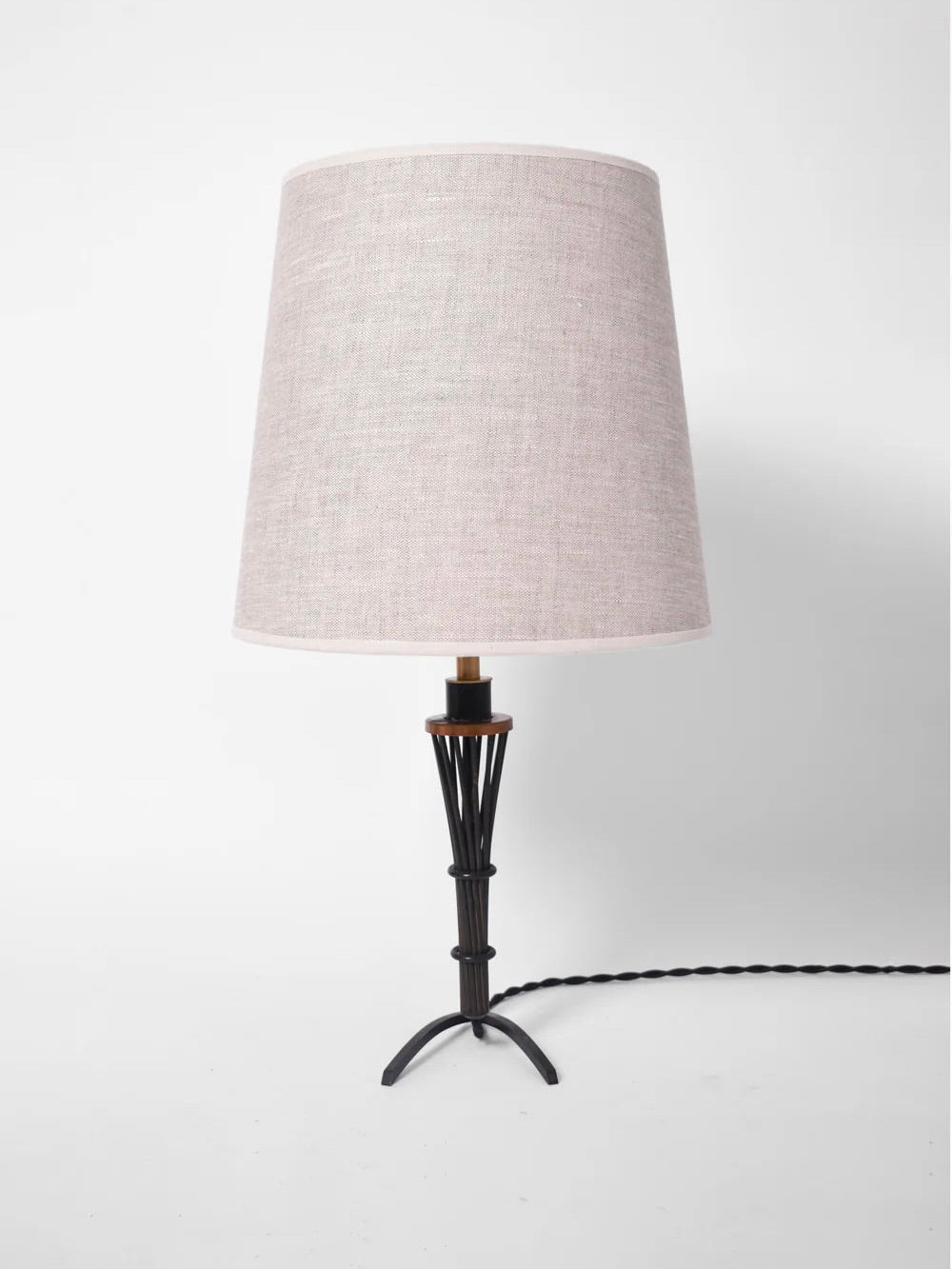 A pair of lamps from Barracuda Interiors, made of iron and copper and inspired by 1940s aesthetics, display a vintage charm with their visible power cords. They feature a tripod base and beige fabric lampshade on a white surface.