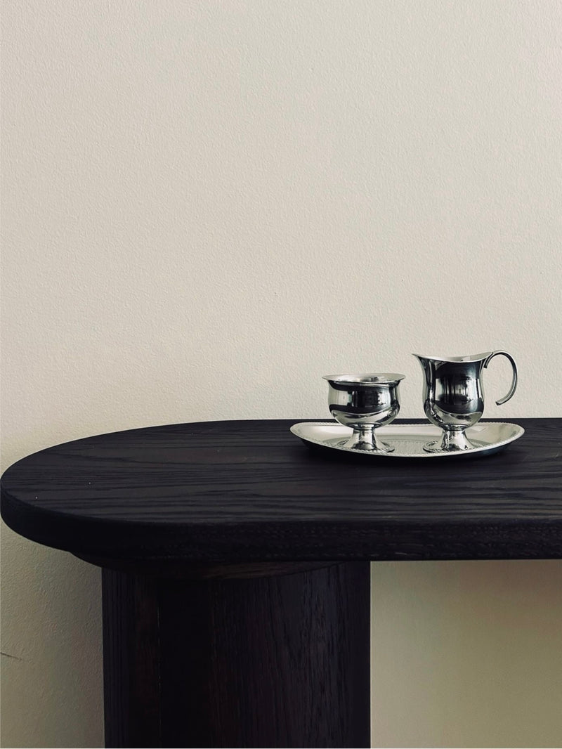 A minimalist scene featuring the Vintage Alessi Sugar & Creamer Set by Flock Studio, with two silver cups and a matching creamer elegantly arranged on a stainless steel tray. This simple and elegant composition rests on a dark wooden table against a plain light wall.
