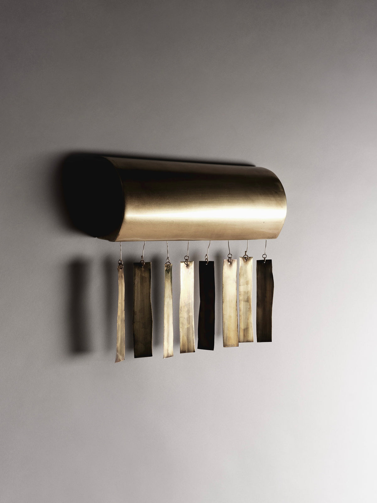 The Quimbaya Sconce by Ombia Studio is a modern wall-mounted sculpture featuring a horizontal, cylindrical brass tube and handmade vertical metal strips that cast soft shadows on a neutral background, echoing minimalist and Quimbaya artistry.