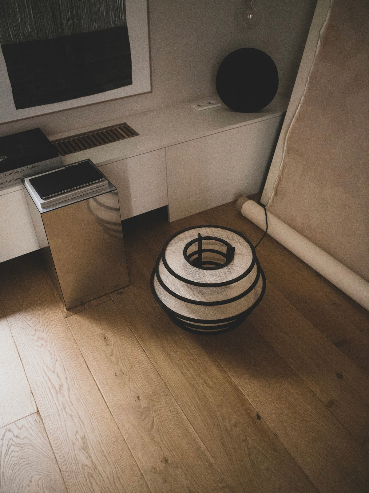 A modern, minimalist room with a wooden floor showcases ELIURPI's Lamp I, featuring black metal accents casting ambient light. A white cabinet holds books and a picture frame, positioned next to a mirrored side table. A rolled canvas leans against the wall, enhancing the handcrafted aesthetic.