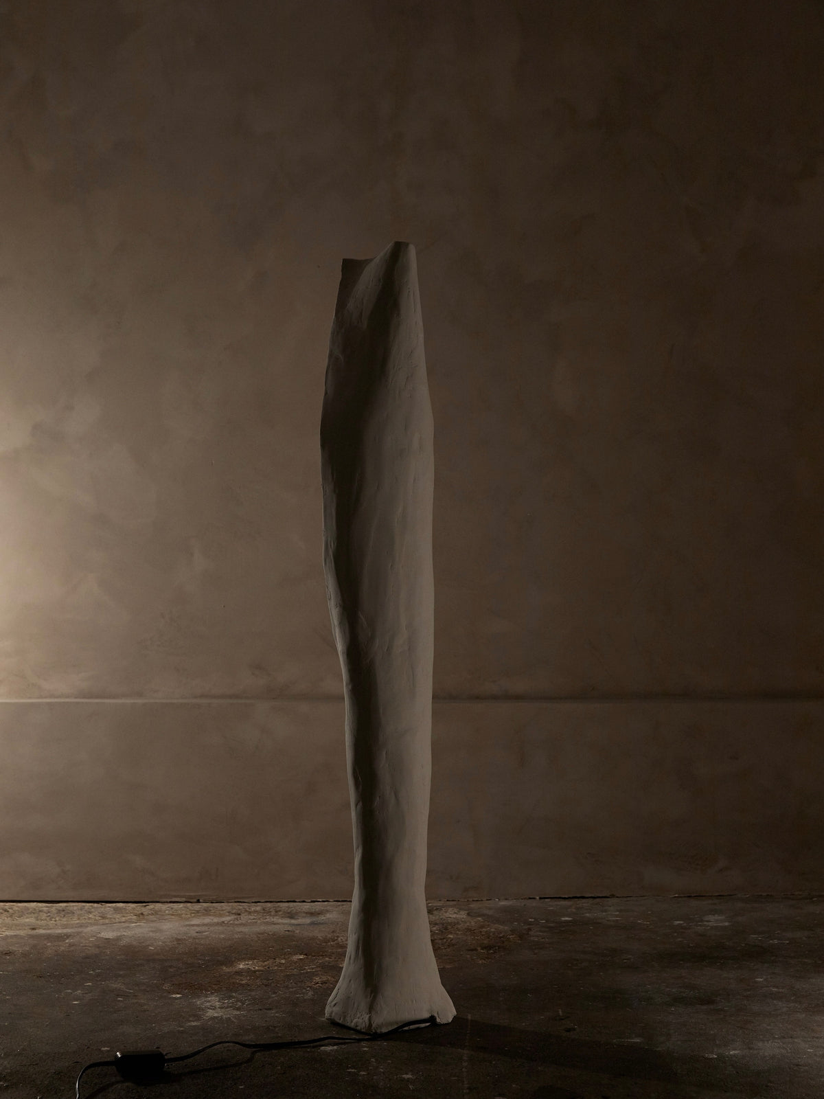In a dim room with textured walls stands the Alcove Floor Lamp by Studio Tristan Kallas—a tall, slender abstract sculpture with an organic shape, gleaming subtly on its smooth white surface as a cable extends from its base.