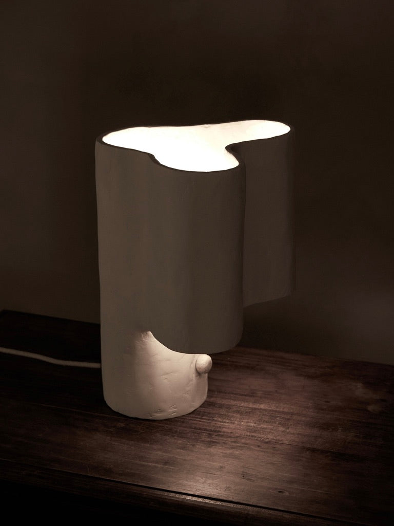 A Cantilever Freeform Table Lamp by Studio Tristan Kallas rests on a wooden surface, featuring a sculptural design with an abstract handcrafted shade casting a soft glow for a warm ambiance. Its smooth cylindrical base blends art and functionality.